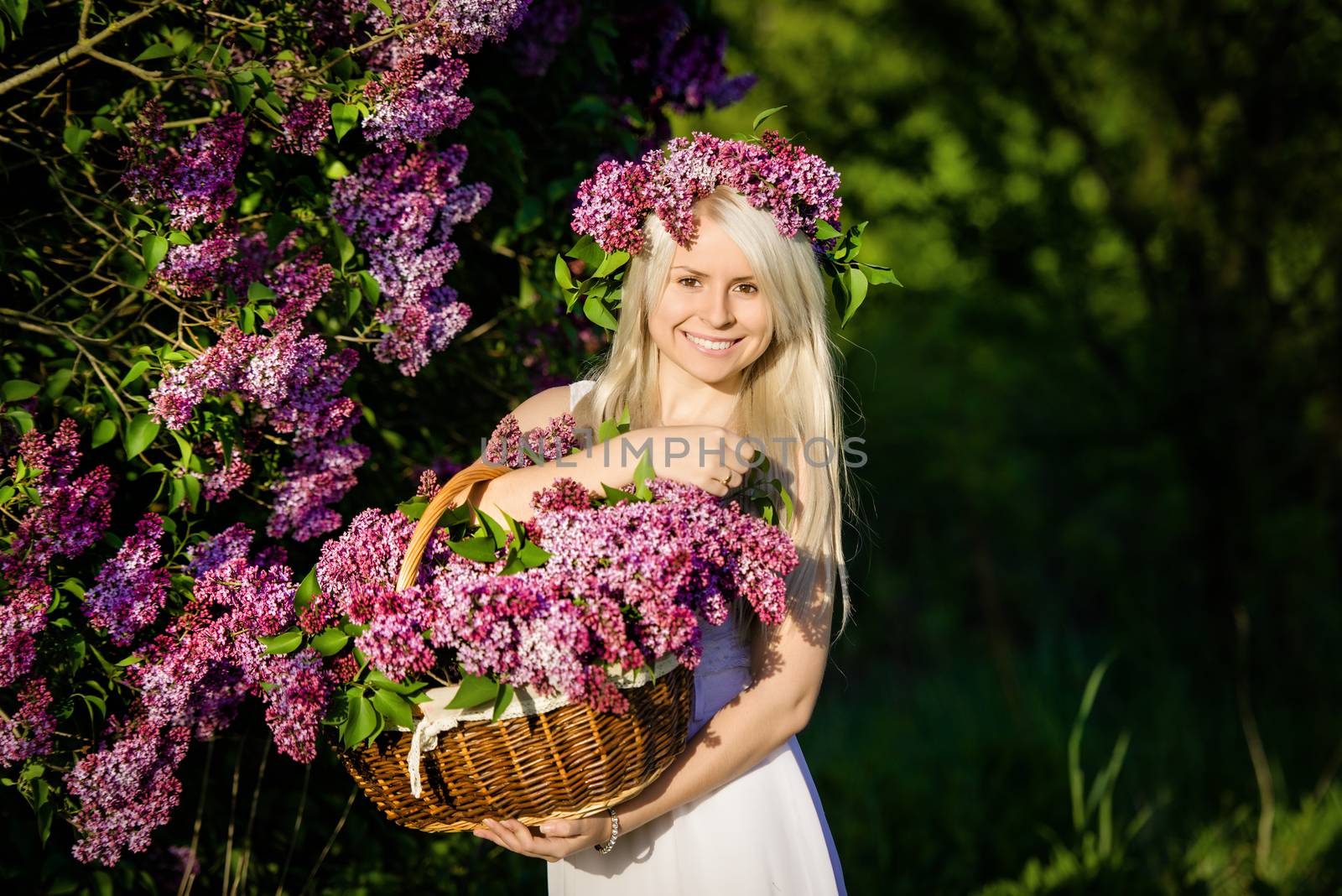 Beautiful smiling young blonde woman is wearing wreath and bouquet lilac flowers in basket, sunset, white dress