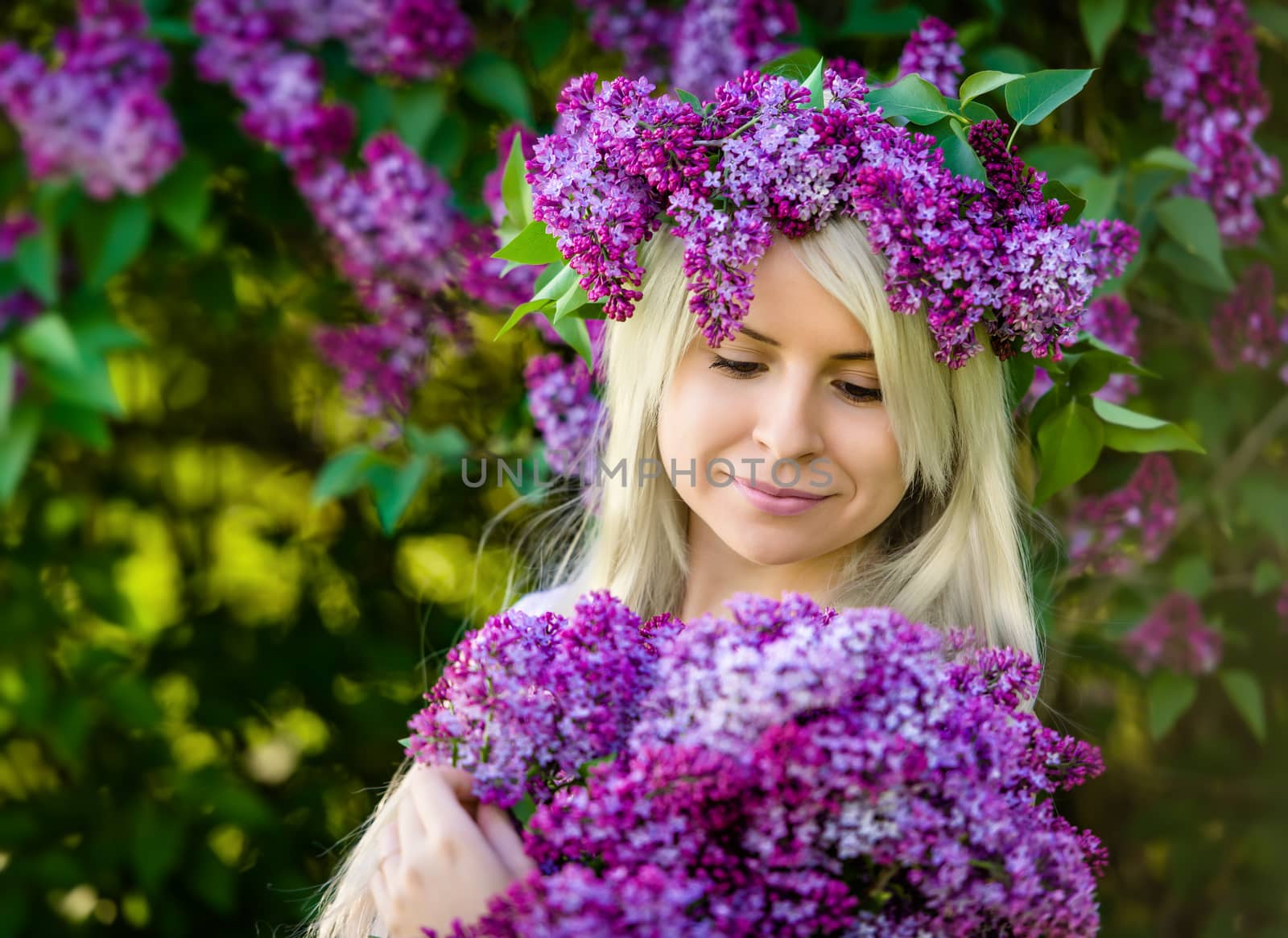 Beautiful smiling young blonde woman is wearing wreath and bouquet of lilac flowers. Outdoor portrait