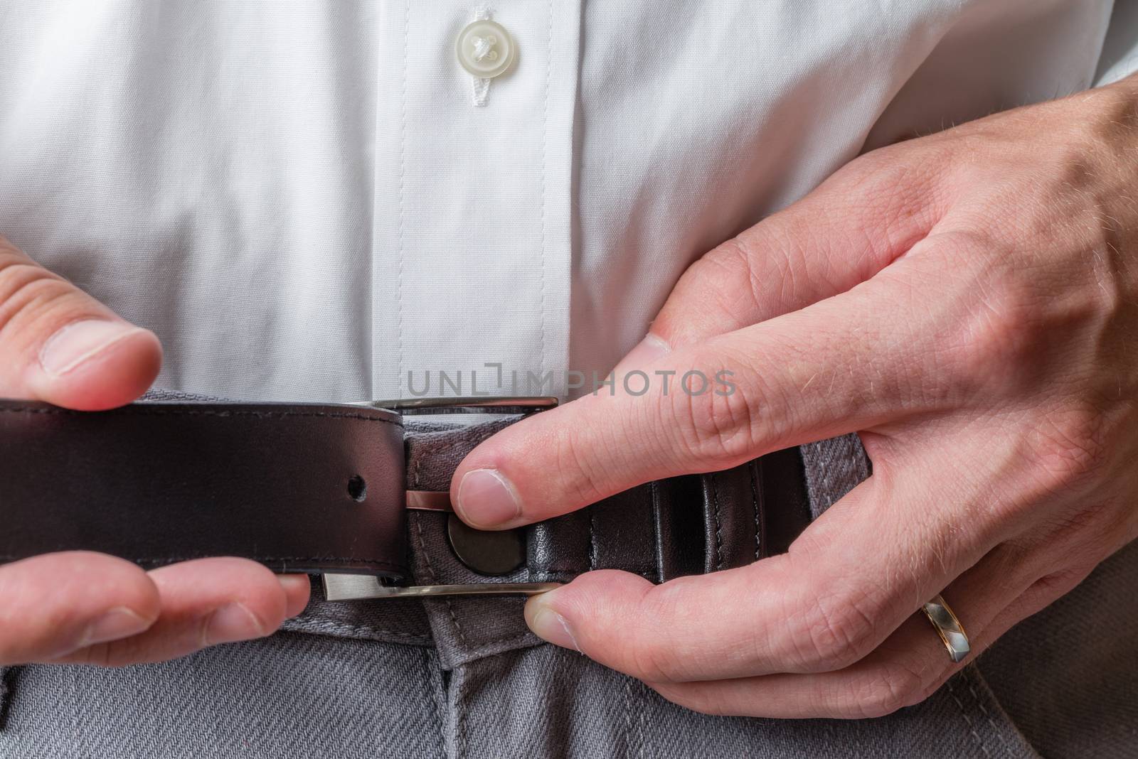 A man wearing grey pants and a white button up shirt buckling his belt.