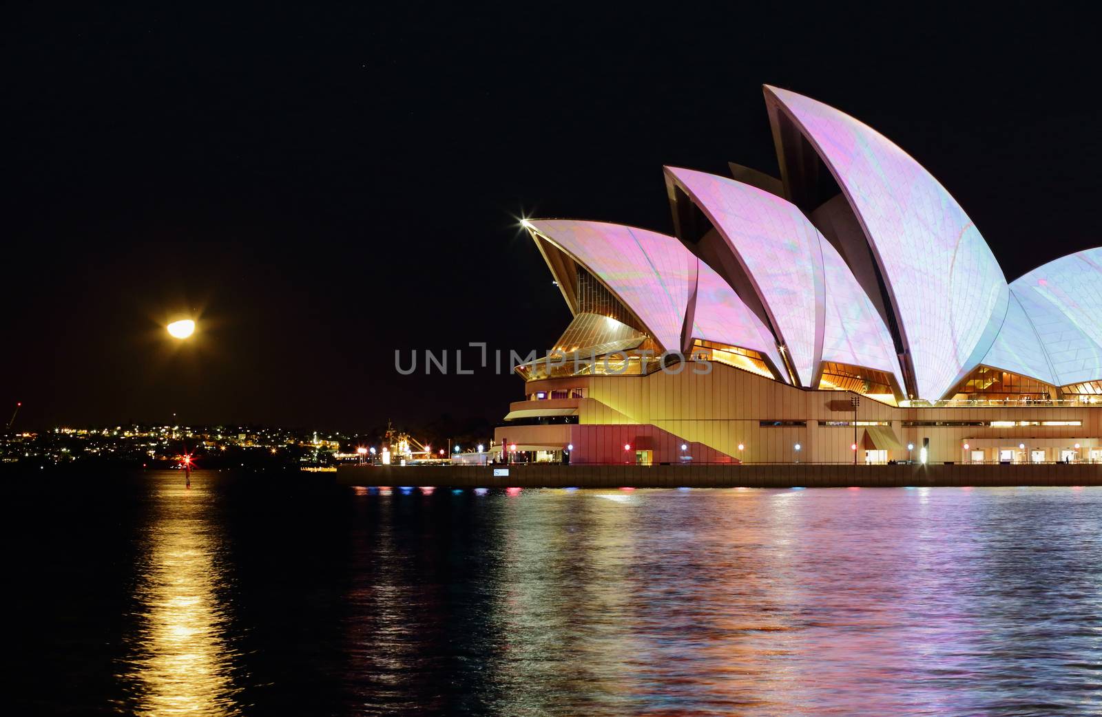 SYDNEY, AUSTRALIA - JUNE 8, 2015;  Sydney Opera House in a myriad of colours and patterns and reflections in the Sydney Harbour waters.  The moon rising in the far east.