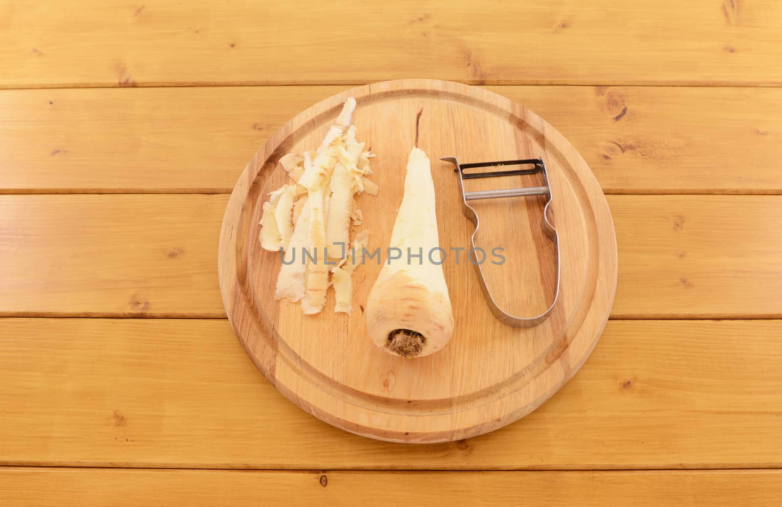 Raw parsnip being peeled with vegetable peeler on a wooden chopping board