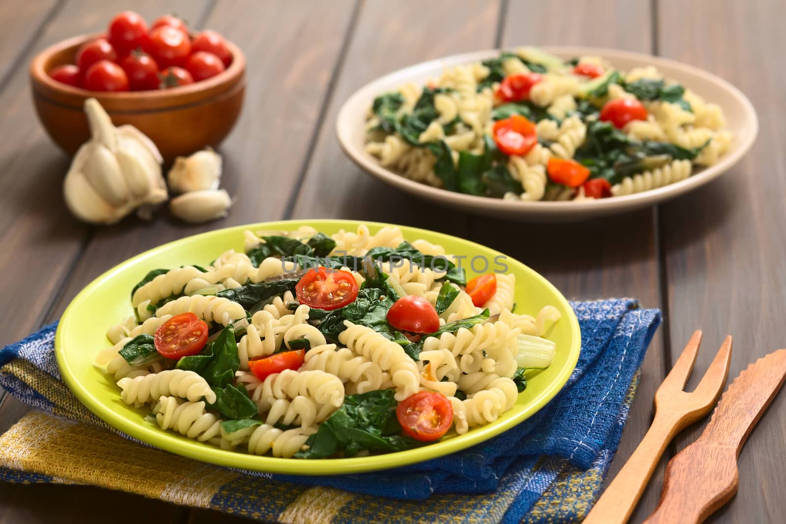 Two plates of fusilli pasta with chard leaves (lat. Beta vulgaris) and cherry tomatoes, photographed on dark wood with natural light (Selective Focus, Focus in the middle of the first dish)