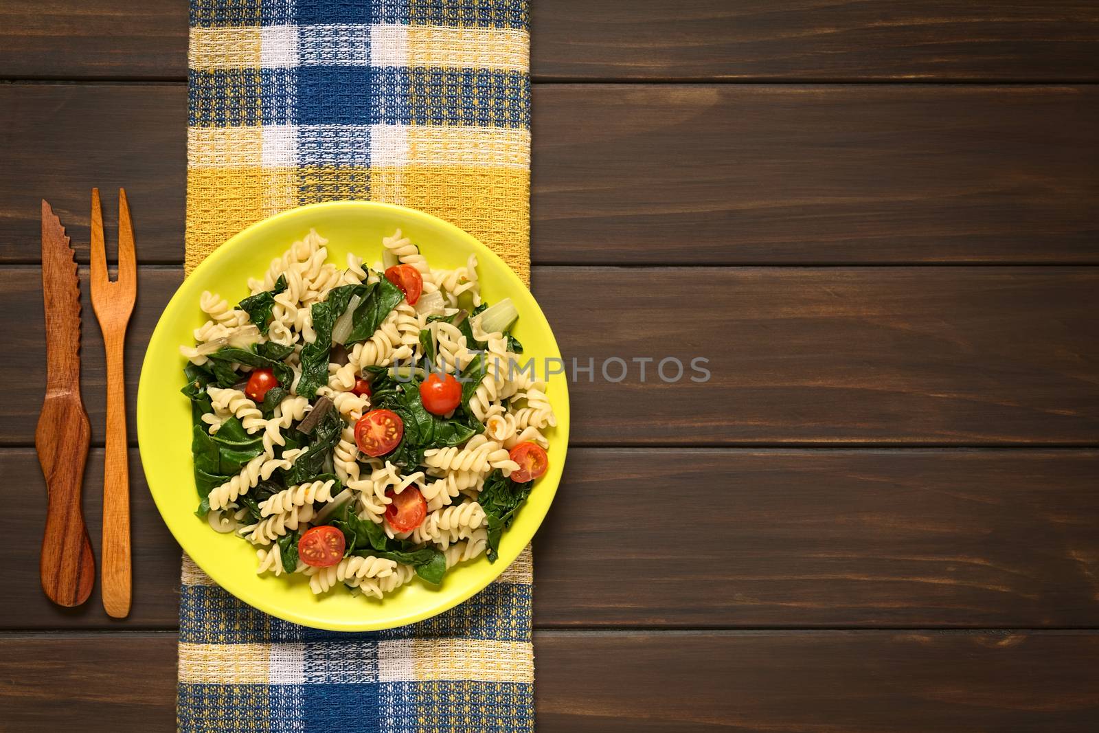 Overhead shot of fusilli pasta with chard leaves (lat. Beta vulgaris) and cherry tomatoes served on plate with wooden fork and knife on the side, photographed on dark wood with natural light