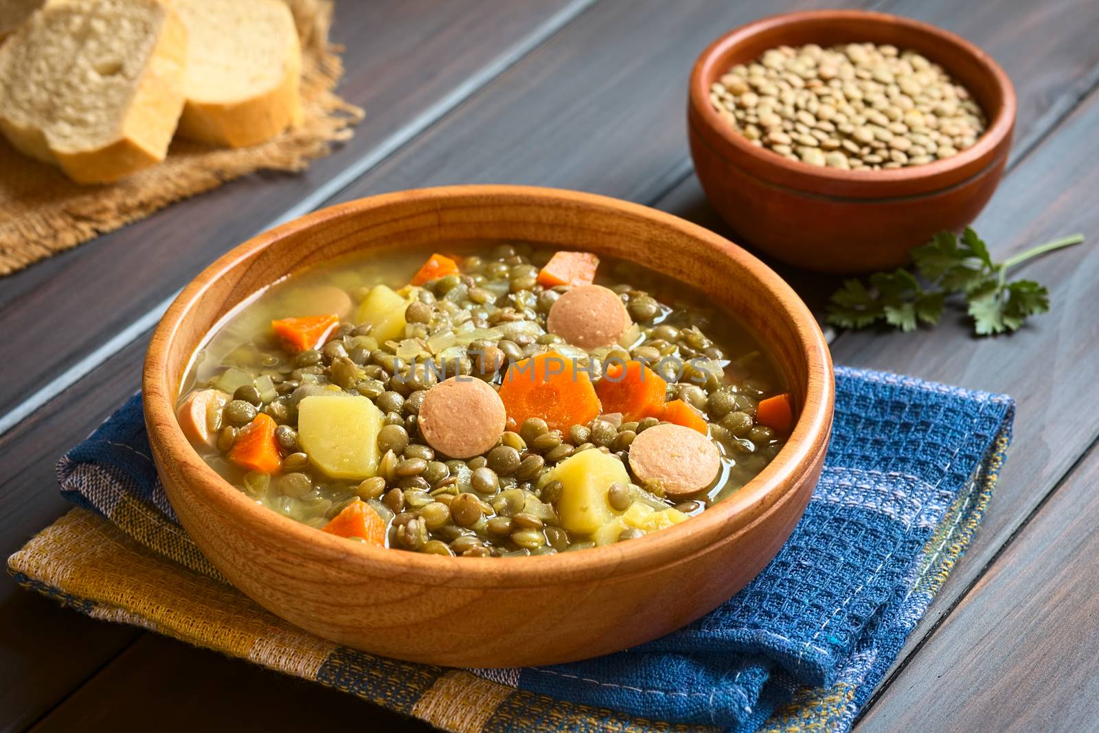 Wooden bowl of lentil soup made with potato, carrot, onion and sausage slices, with a small bowl of raw lentils in the back, photographed on dark wood with natural light (Selective Focus, Focus on the middle of the soup)