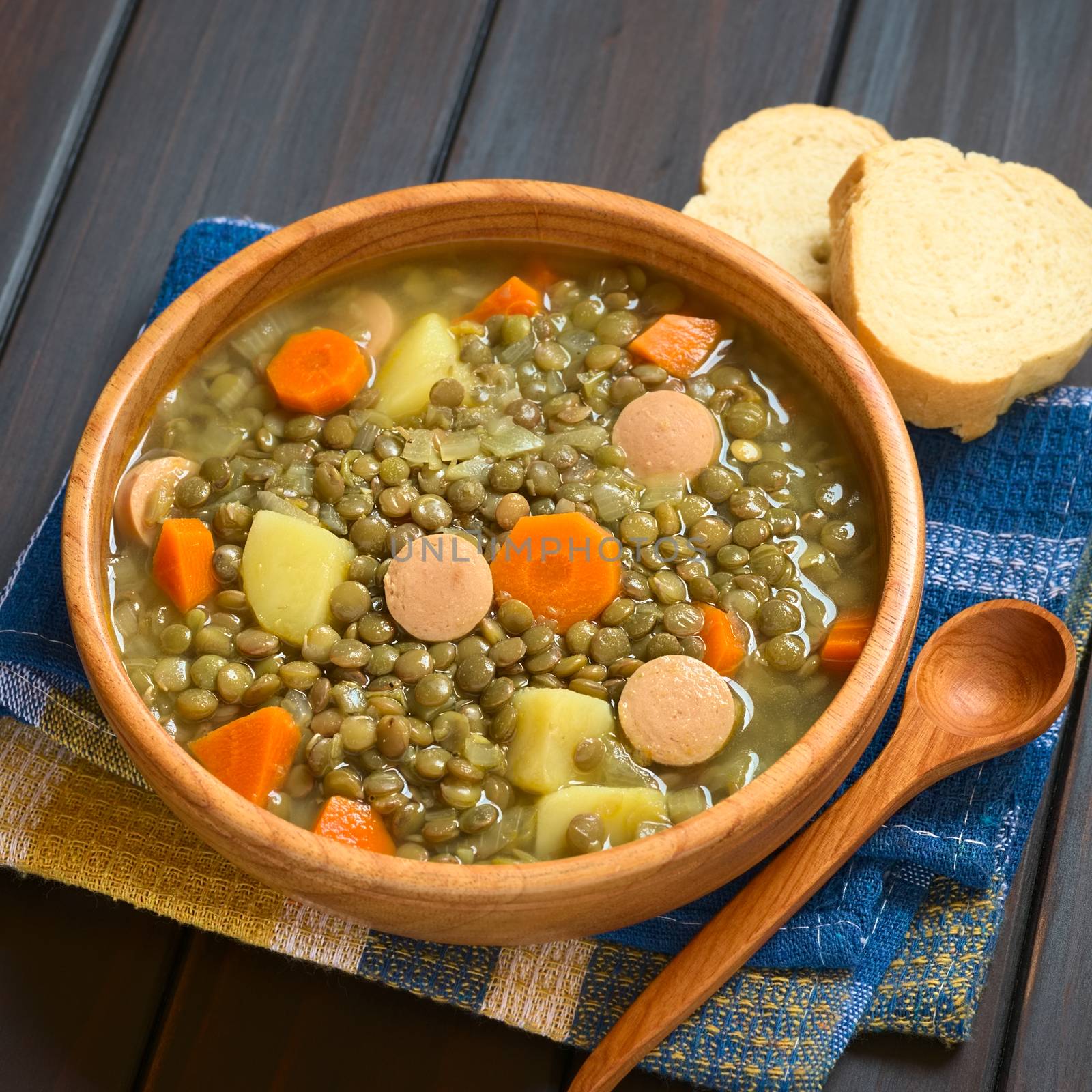 Wooden bowl of lentil soup made with potato, carrot, onion and sausage slices, photographed on dark wood with natural light (Selective Focus, Focus on the middle of the soup)