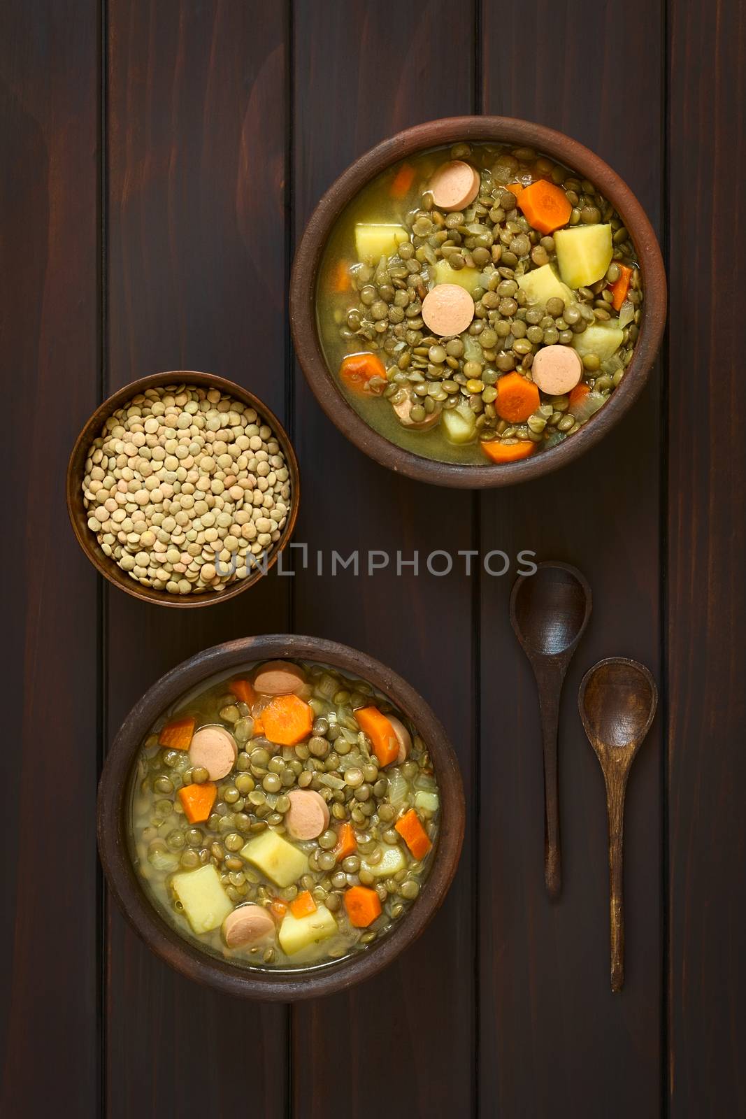 Overhead shot of two rustic bowls of lentil soup made with potato, carrot, onion and sausage slices, with a small bowl of raw lentils and wooden spoons on the side, photographed on dark wood with natural light
