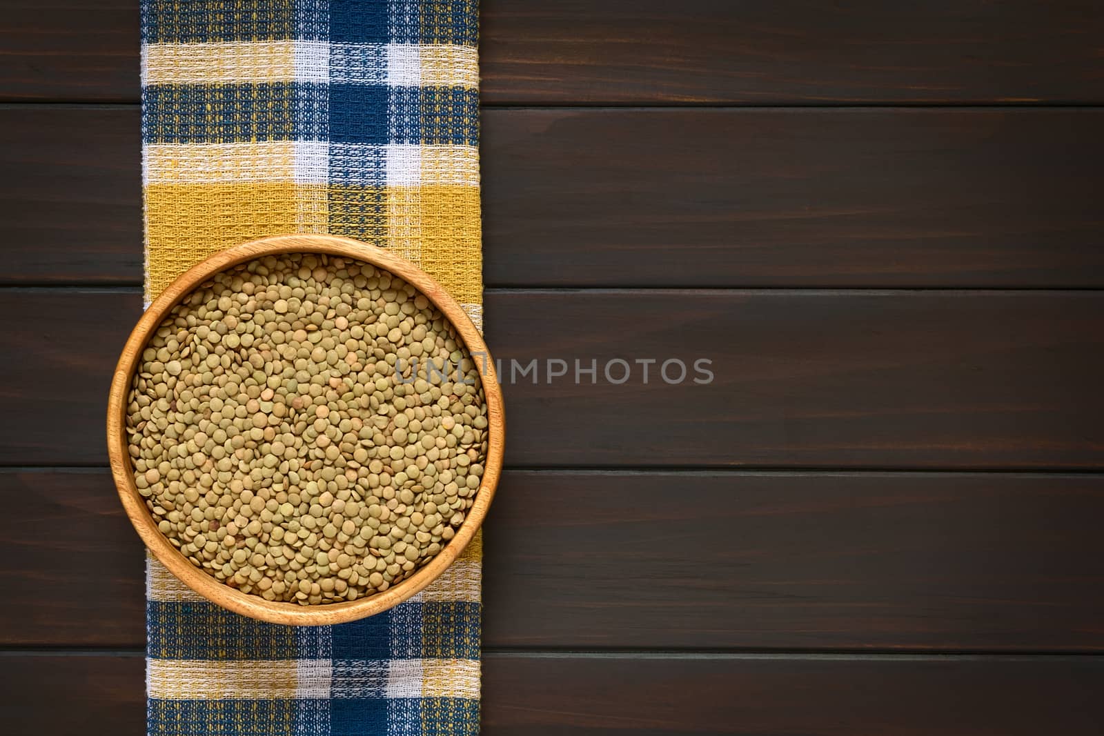 Overhead shot of raw lentils (lat. Lens culinaris) in wooden bowl, photographed on kitchen towel on dark wood with natural light