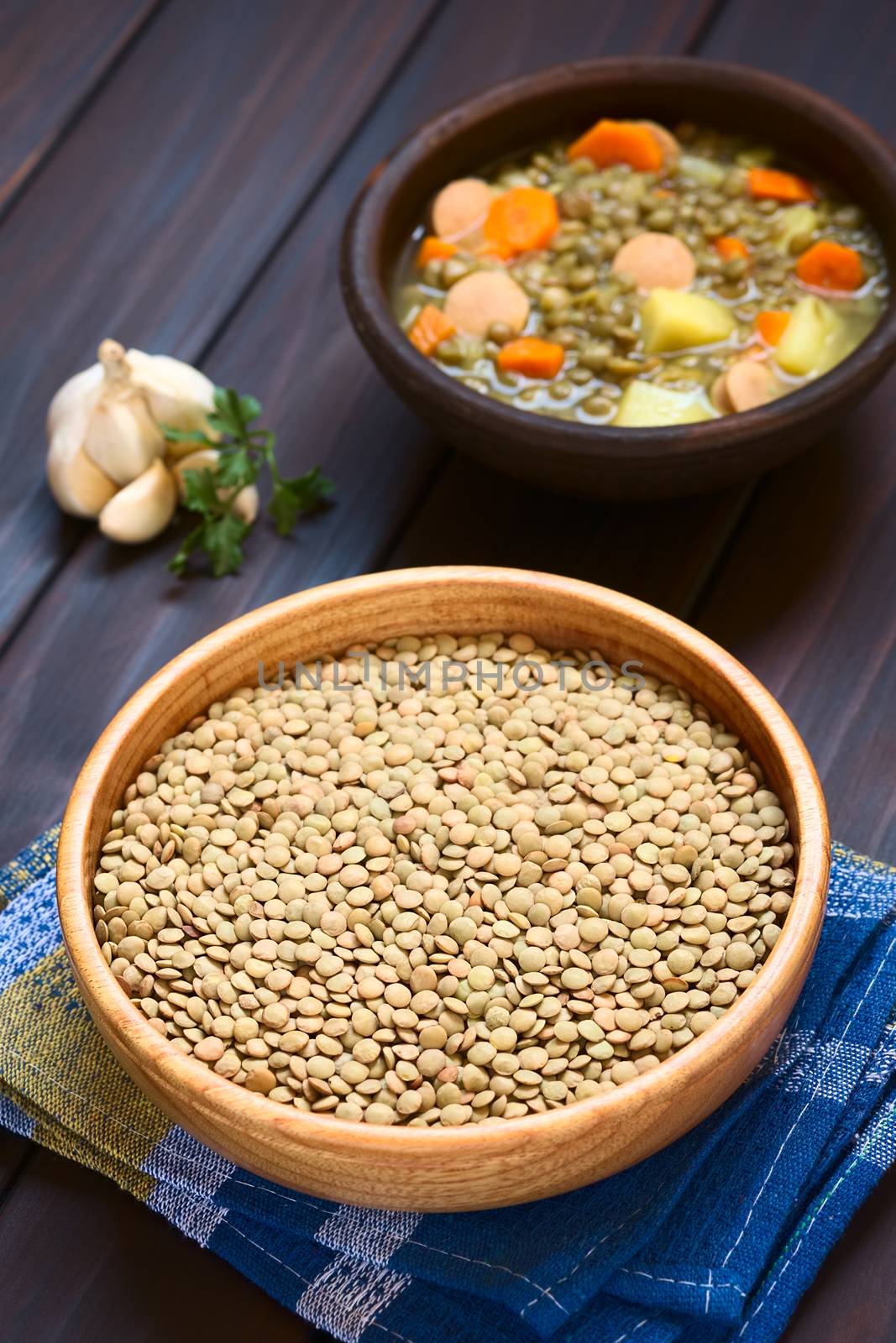 Raw lentils (lat. Lens culinaris) in wooden bowl with a rustic bowl of lentil soup (made with lentil, carrot, potato, sausage) in the back, photographed on dark wood with natural light (Selective Focus, Focus one third into the raw lentils)