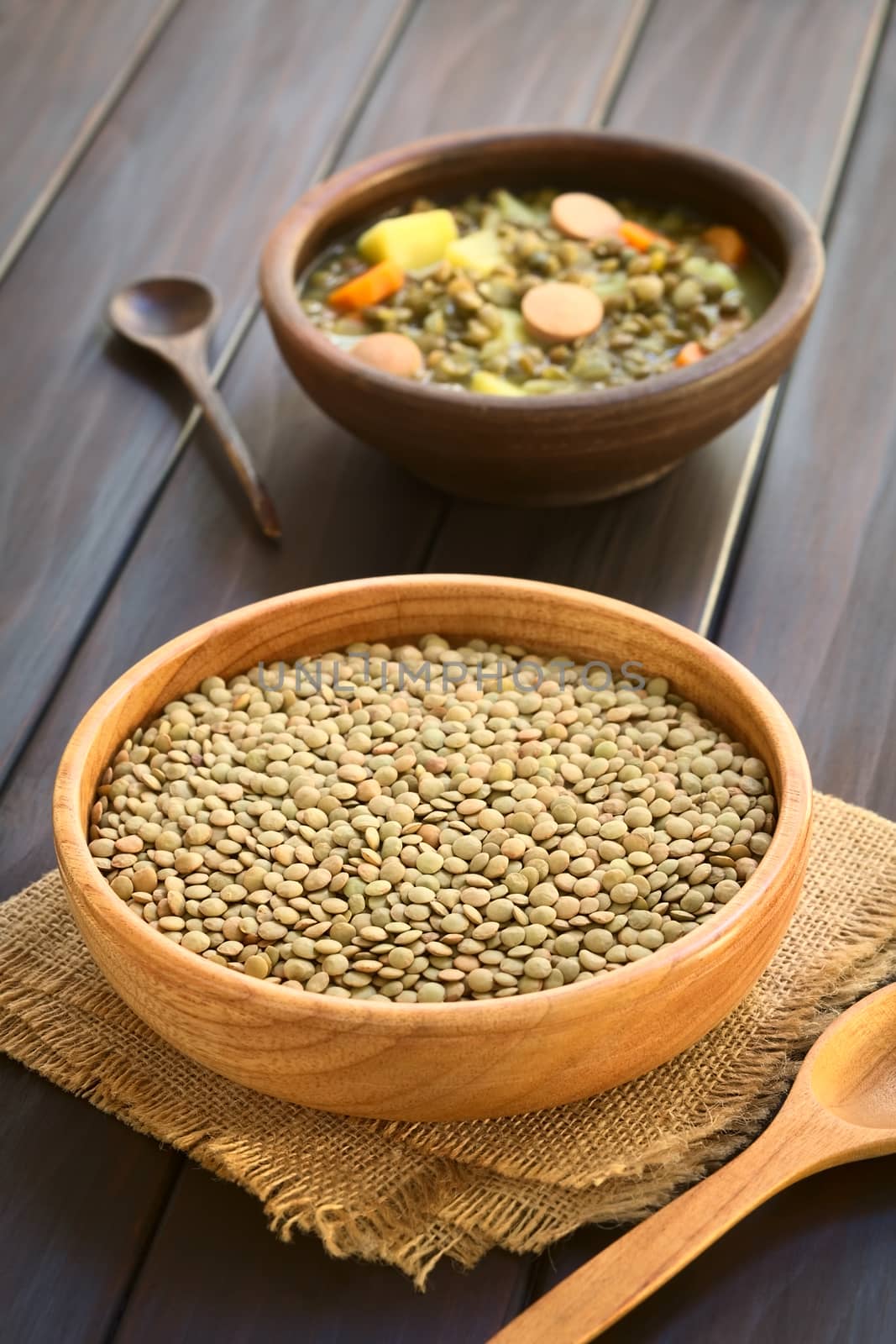 Raw lentils (lat. Lens culinaris) in wooden bowl with a rustic bowl of lentil soup (made with lentil, carrot, potato, sausage) in the back, photographed on dark wood with natural light (Selective Focus, Focus one third into the raw lentils)