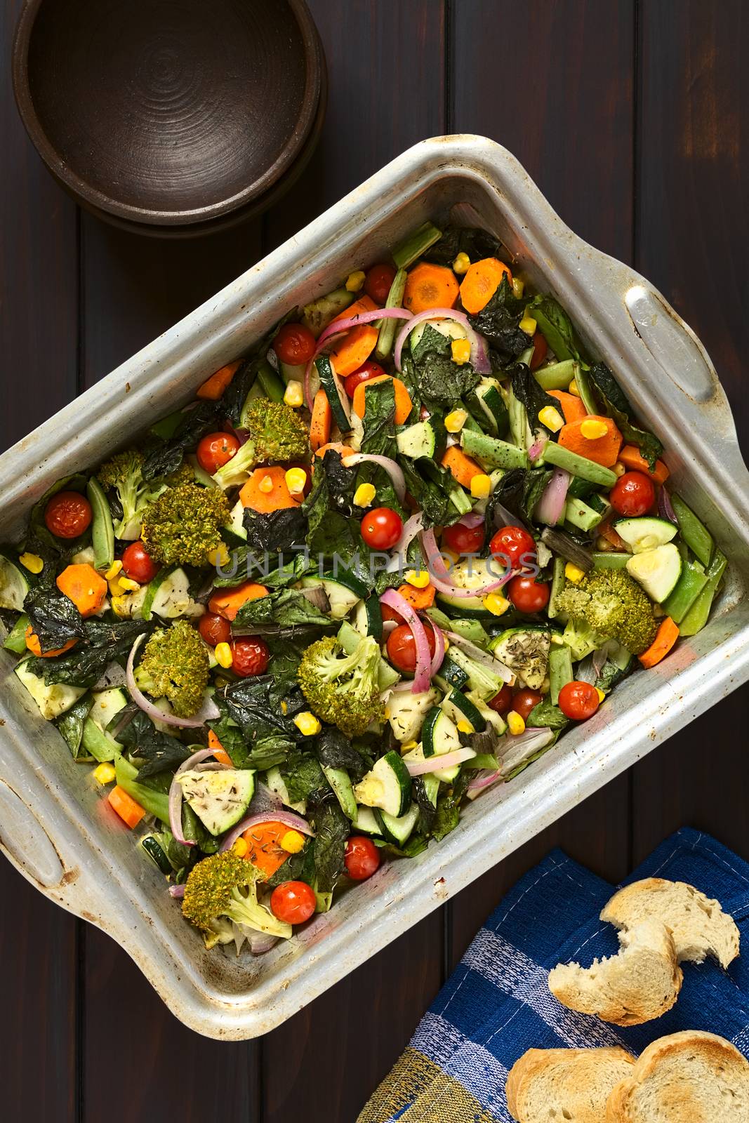 Overhead shot of baked vegetables (zucchini, onion, cherry tomato, broccoli, carrot, sweet corn, green bean, chard) seasoned with thyme in baking dish, photographed on dark wood with natural light