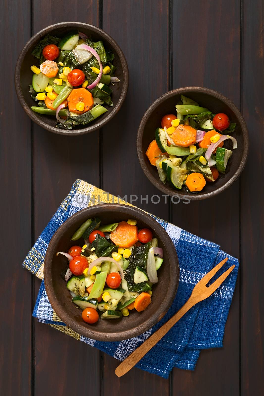 Overhead shot of three rustic bowls of baked vegetables (zucchini, onion, cherry tomato, broccoli, carrot, sweet corn, green bean, chard) seasoned with thyme, photographed on dark wood with natural light