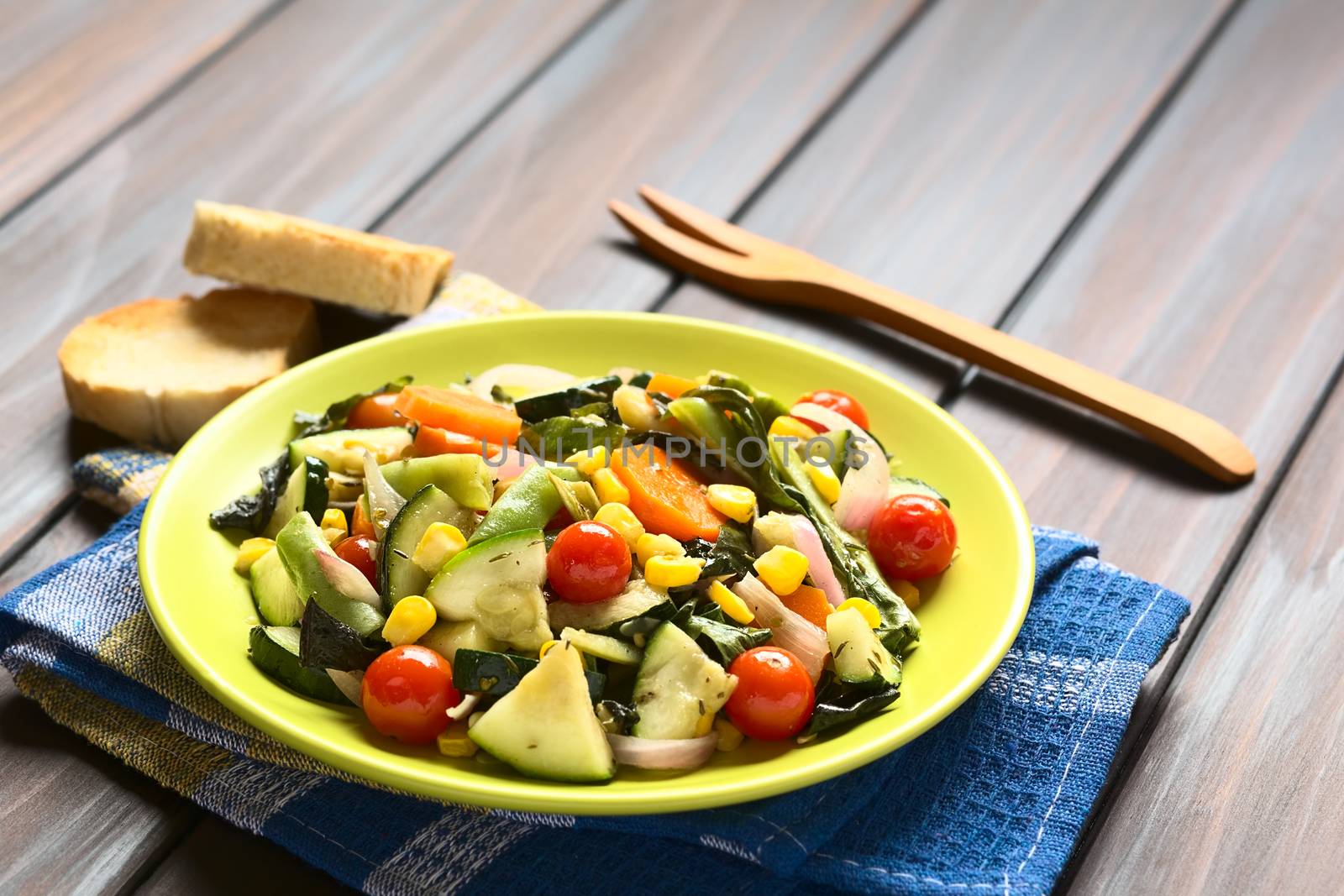 Baked vegetables (zucchini, onion, cherry tomato, broccoli, carrot, sweet corn, green bean, chard) seasoned with thyme served on plate, photographed on dark wood with natural light (Selective Focus, Focus one third into the dish)