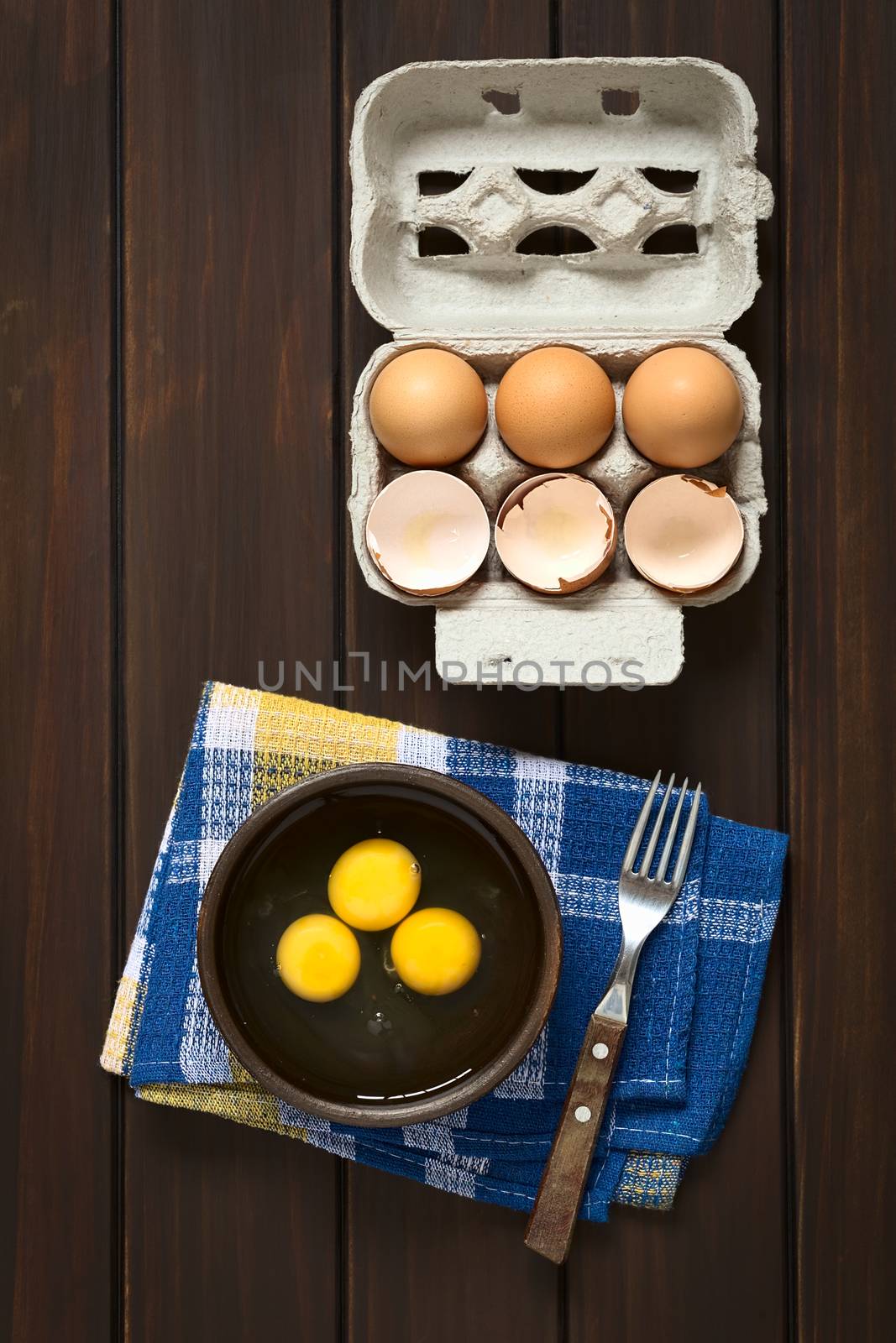 Overhead shot of three raw eggs in rustic bowl with fork on the side and egg box with eggs and eggshells above, photographed on dark wood with natural light