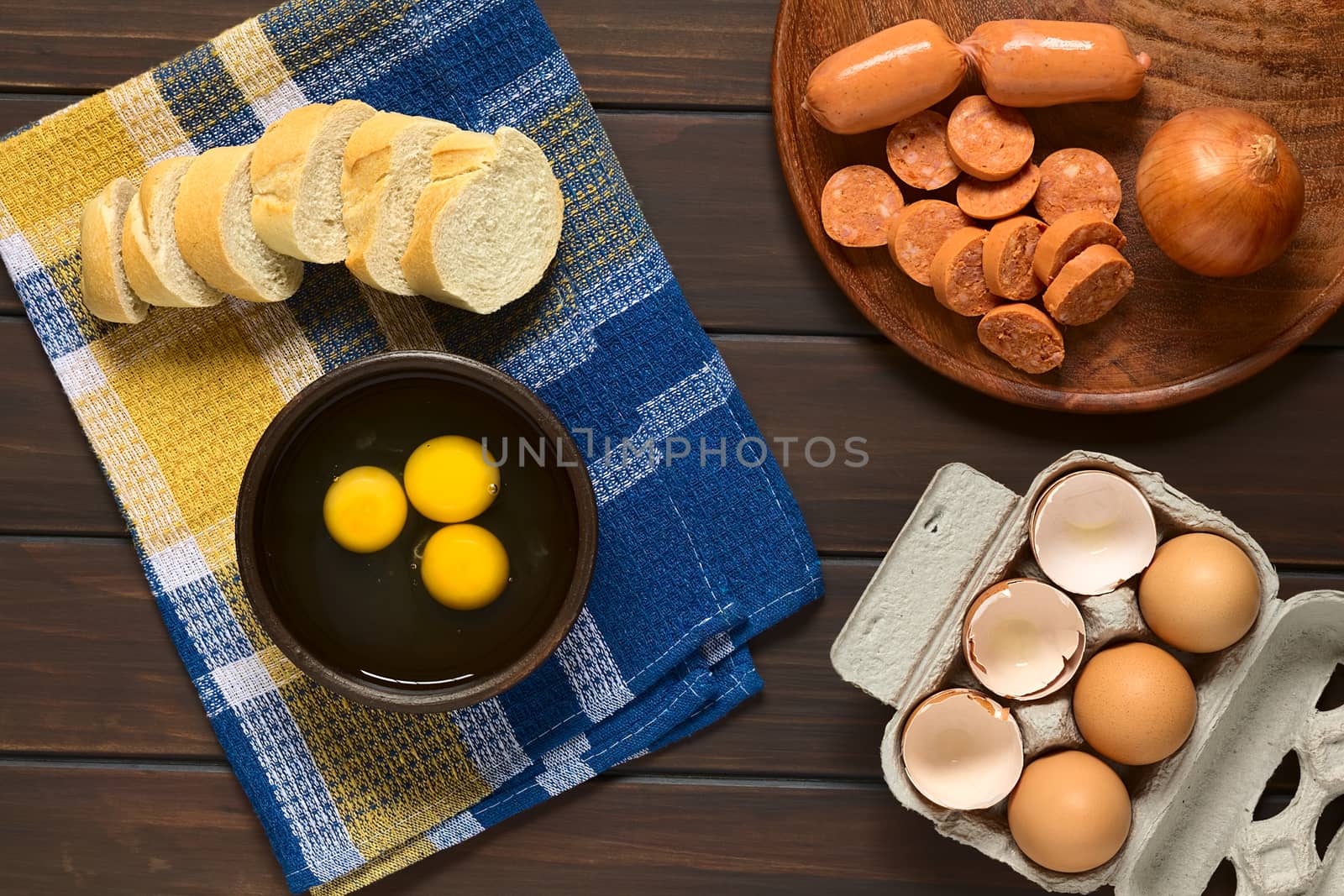 Overhead shot of three raw eggs in rustic bowl with baguette slices, sausage, onion and egg box with eggs and eggshells, photographed on dark wood with natural light