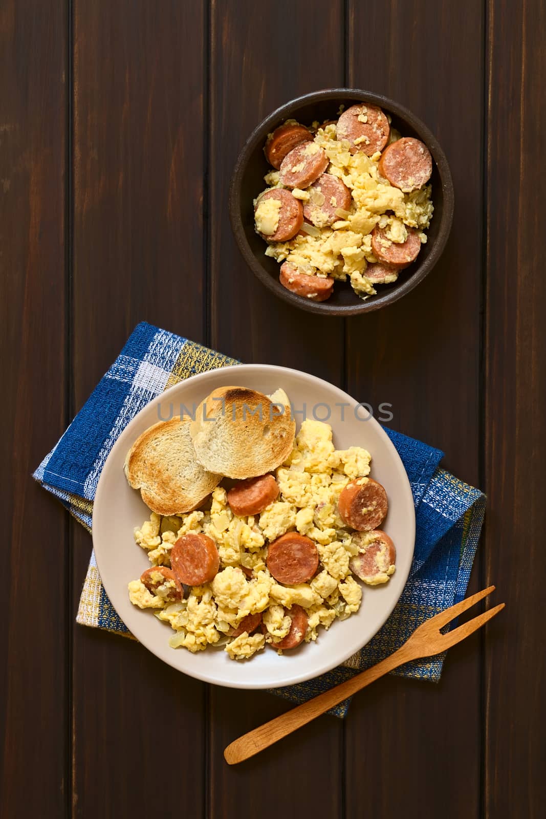 Overhead shot of scrambled eggs made with chorizo slices and onion on plate with toasted baguette slices, wooden fork on the side, photographed on dark wood with natural light