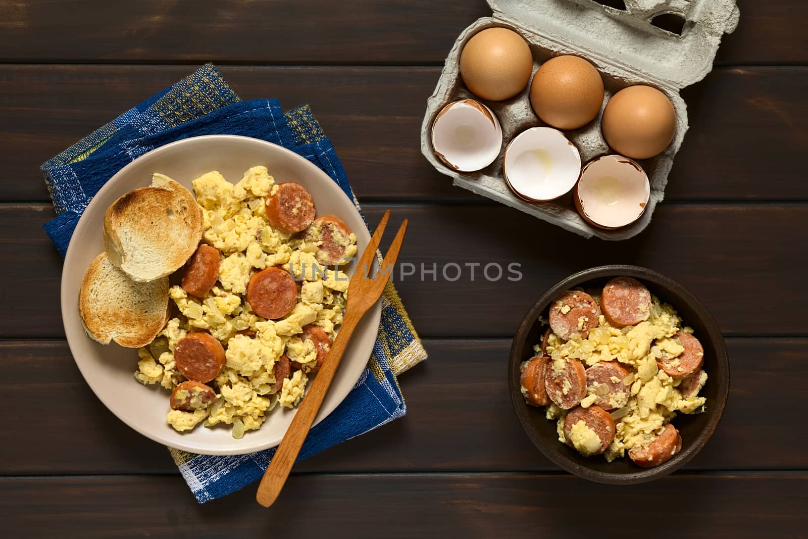 Overhead shot of scrambled eggs made with chorizo slices and onion on plate with toasted baguette slices and a wooden fork, an egg box with eggs and eggshells on the side, photographed on dark wood with natural light