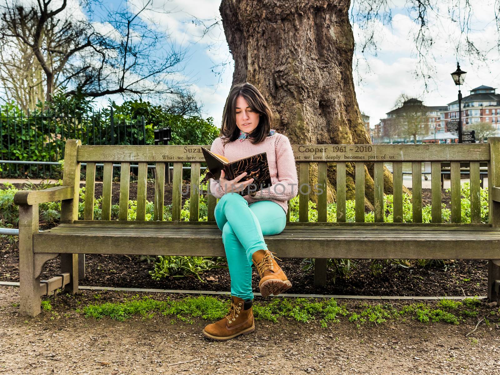 A Casual Young Woman Sitting on a Bench in a Park and Reading Her Book