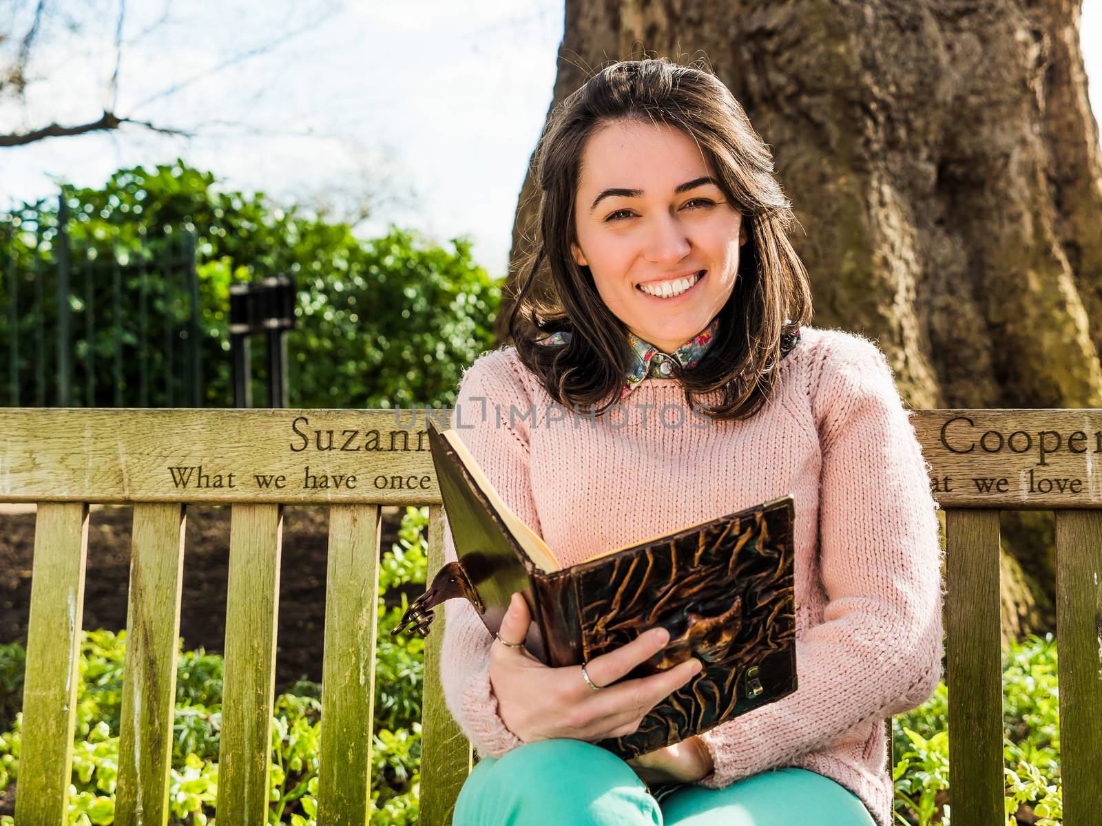 Young Beauty Woman Holding Book and Smiling Seated on Park Bench Looking at the Camera