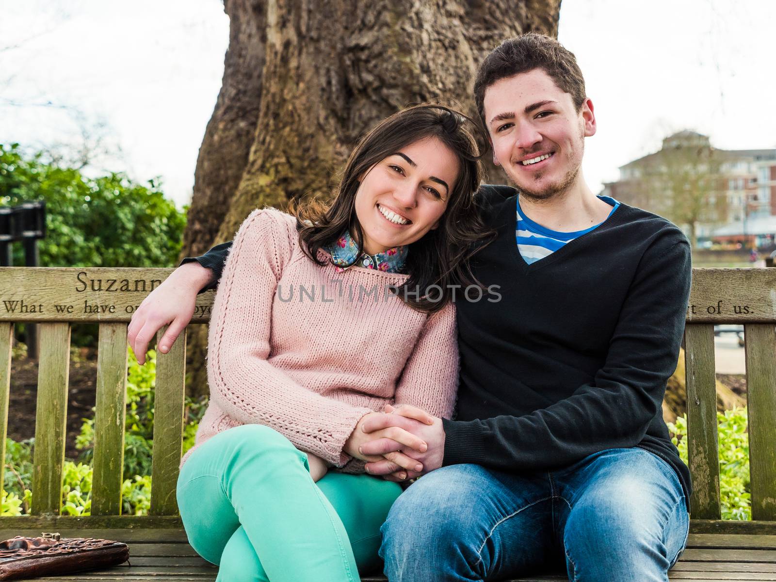 Young Heterosexual Couple Sitting on a Park Bench Enjoying a Beautiful Sunny Day and Smiling Looking at the Camera