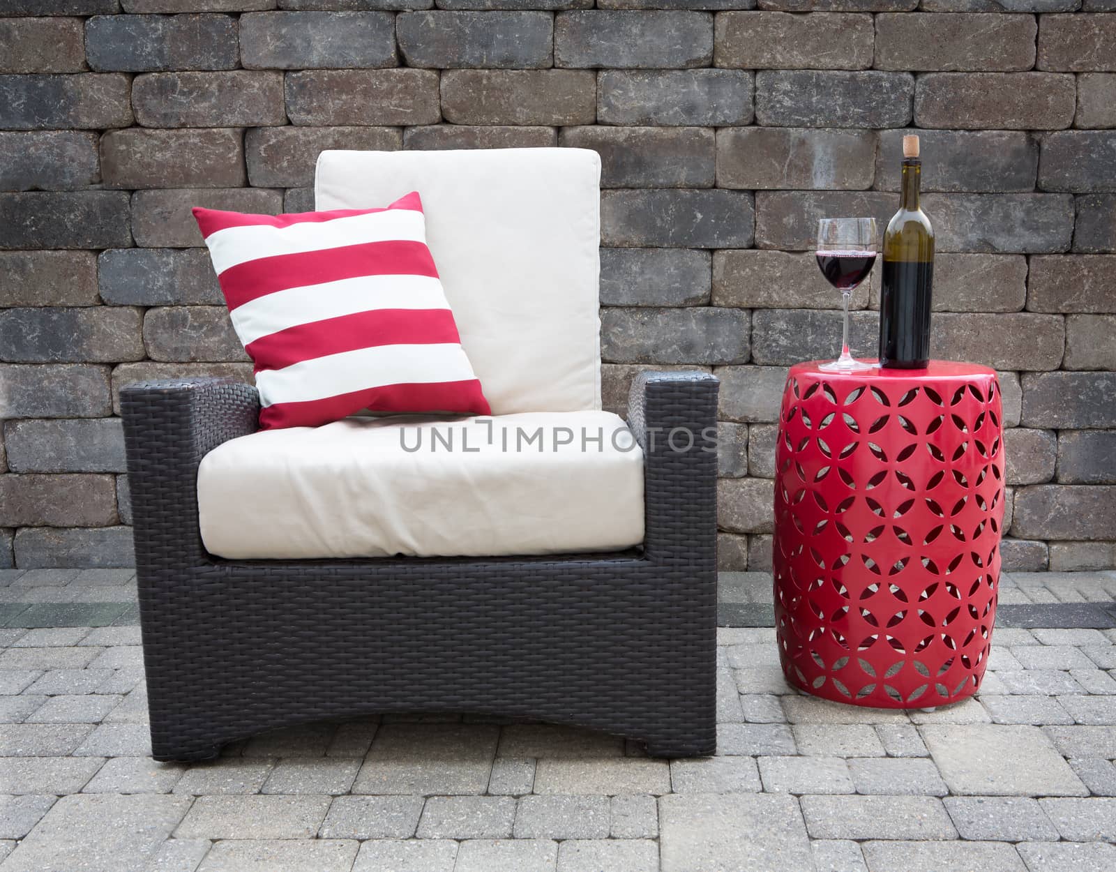 Comfortable Dark Wicker Patio Chair Outfitted with Plush Cushions Beside Modern Red Table with Bottle and Glass of Red Wine on Outdoor Stone Affluent Patio