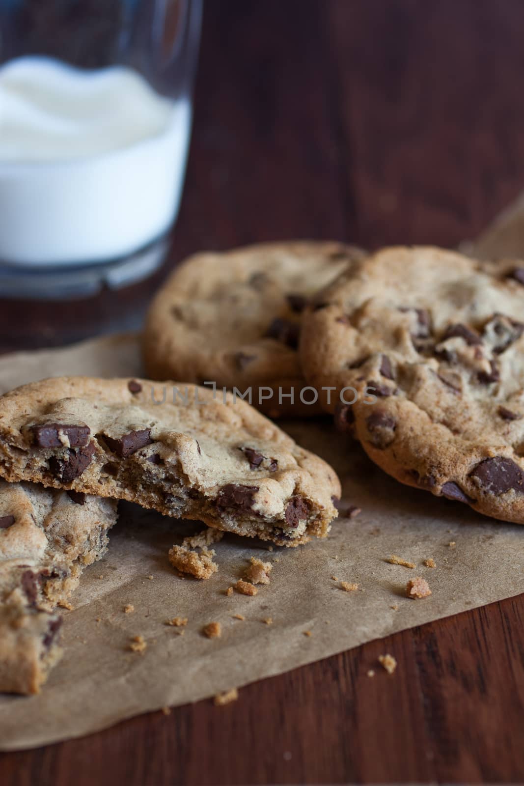 Fresh chocolate chunk cookies on a piece of brown paper  with a glass of miolk on a wooden table.
