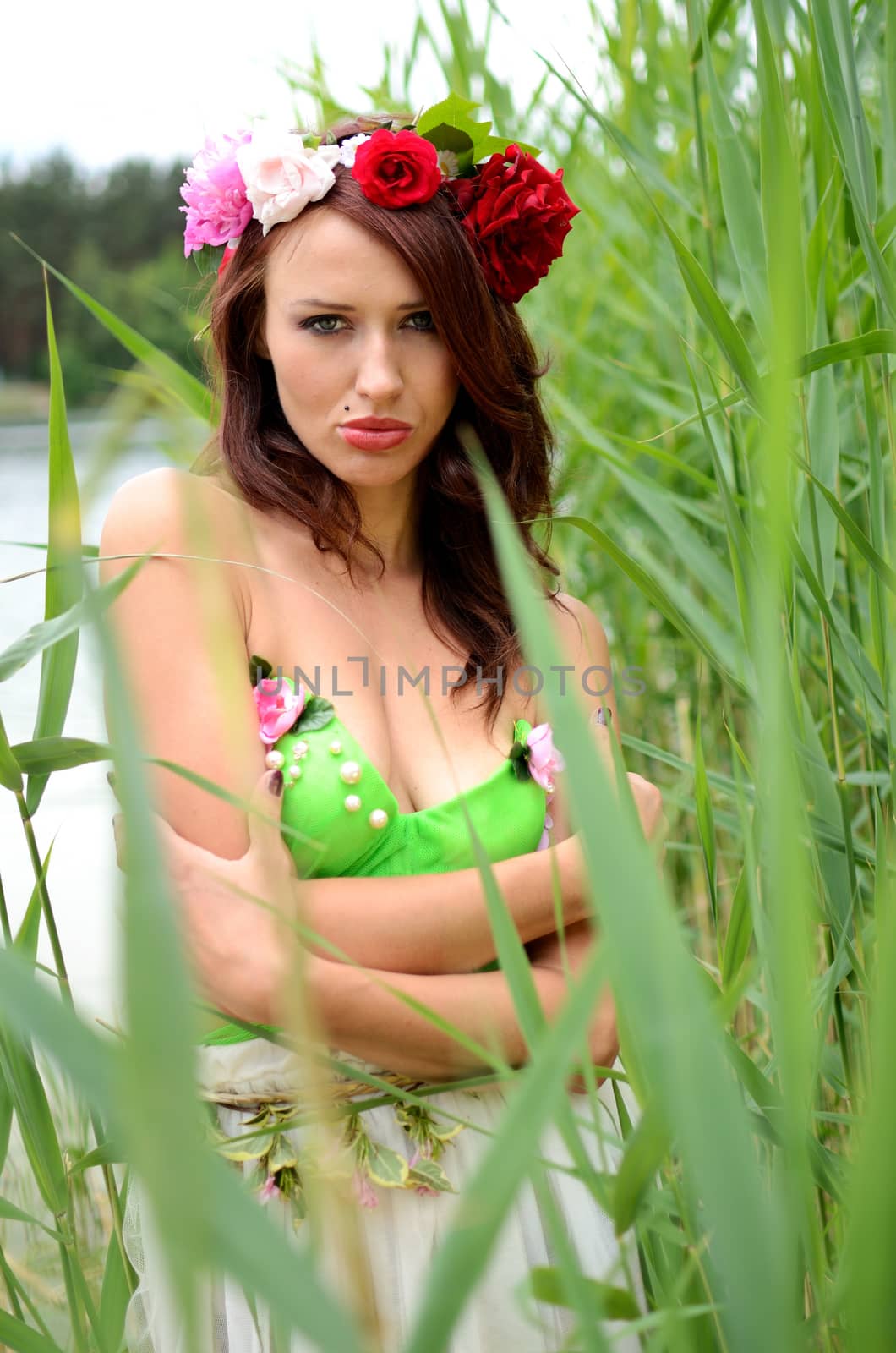 Portrait of female model with chestnut hairs, wearing wreath made of mixed flowers. Woman posing near lake surrounded by reeds.  