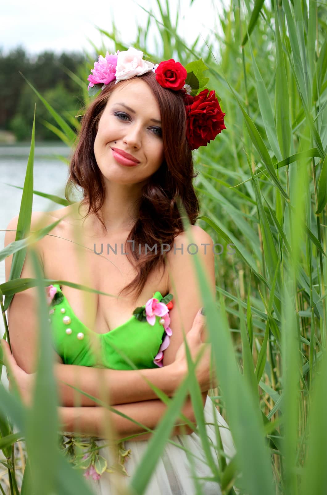 Portrait of female model with chestnut hairs, wearing wreath made of mixed flowers. Woman posing near lake surrounded by reeds.  