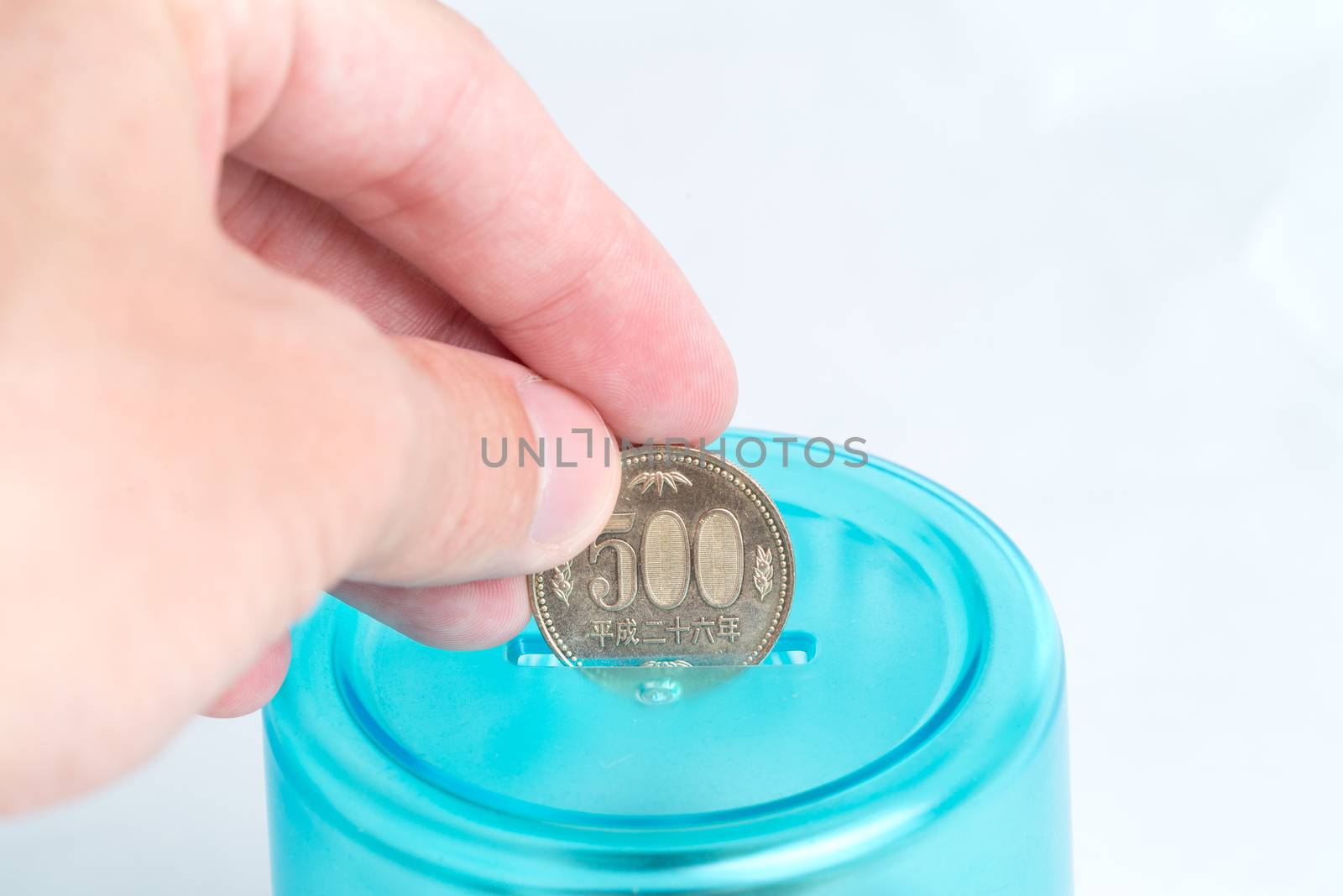 A man's hand putting a 500 Japanese Yen coin into a blue bank with a white background.