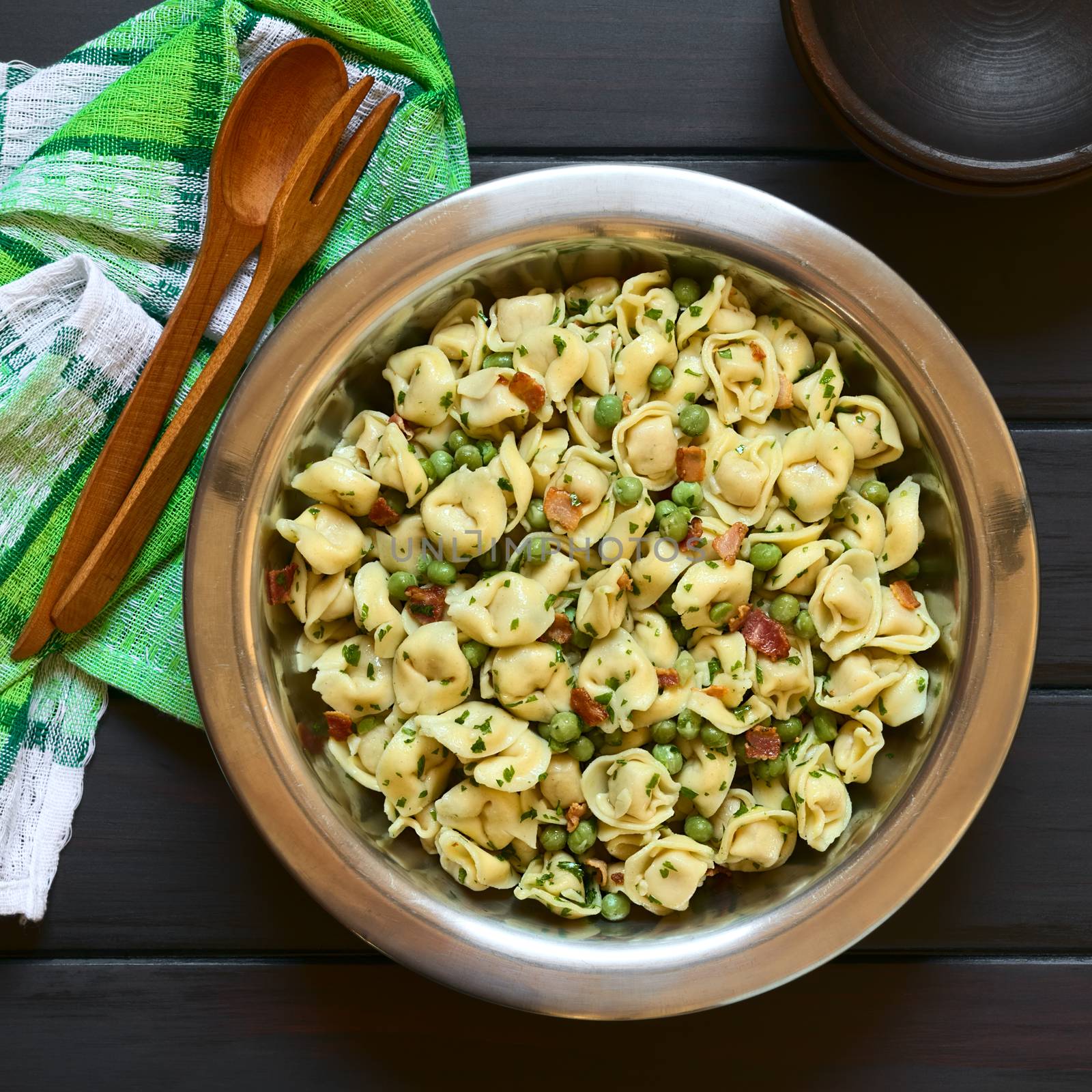 Tortellini salad with green peas, fried bacon and parsley in big salad bowl, with small rustic bowls, wooden spoon and fork on the side, photographed overhead on dark wood with natural light
