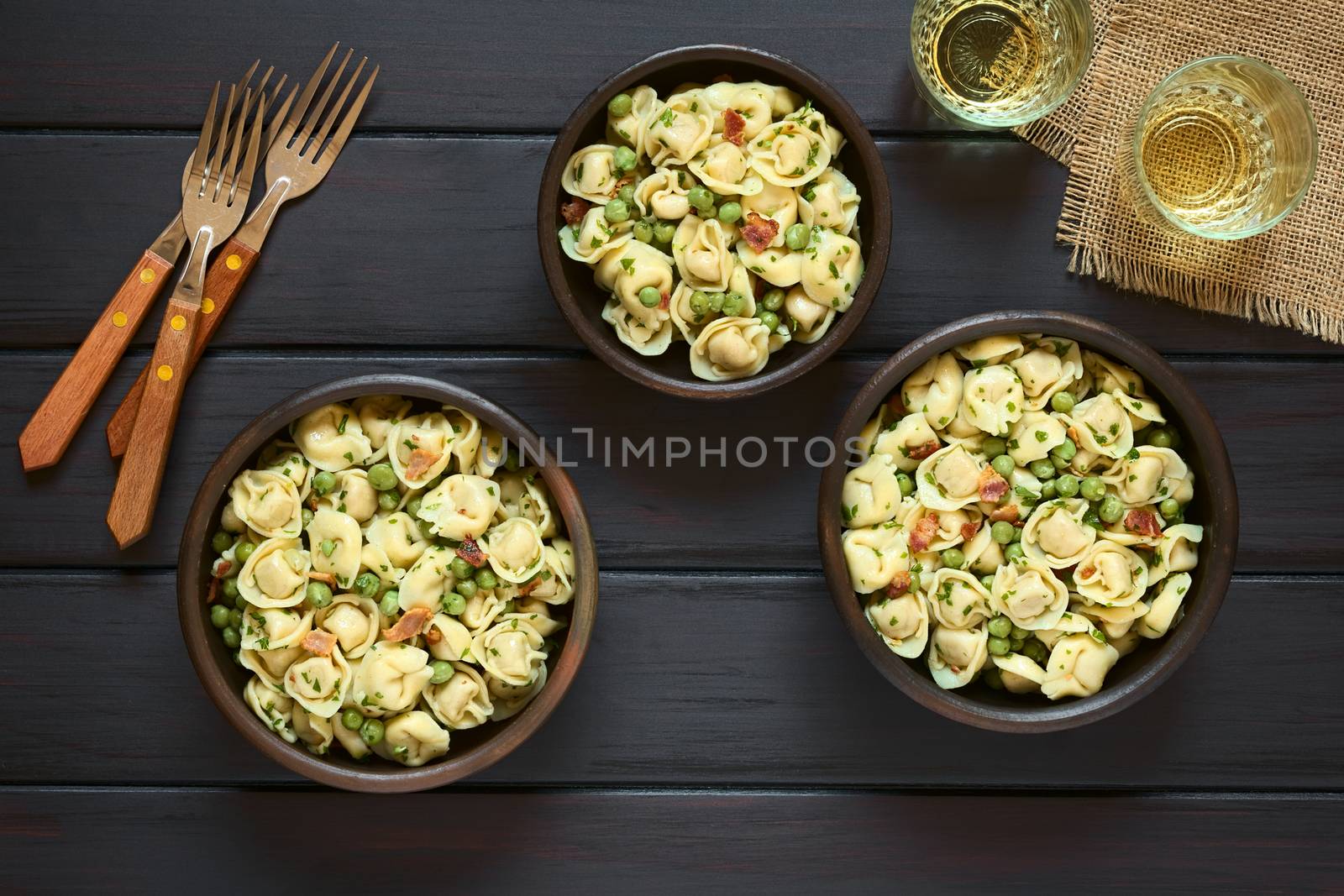 Tortellini salad with green peas, fried bacon and parsley served in rustic bowls, with forks and glasses of white wine on the side, photographed overhead on dark wood with natural light