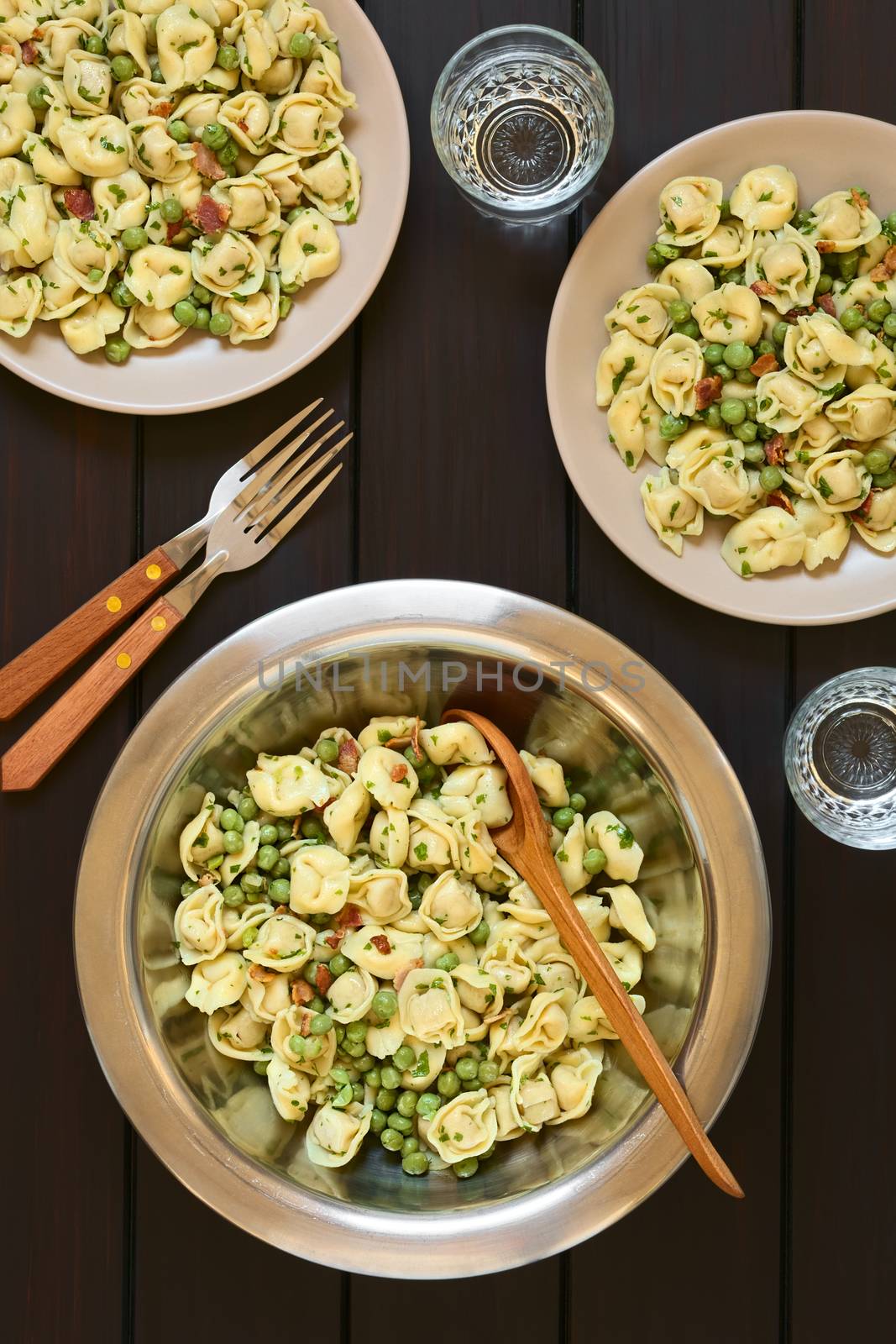 Tortellini salad with green peas, fried bacon and parsley in big salad bowl with spoon to serve, salad served on two plates, forks and glasses, photographed overhead on dark wood with natural light
