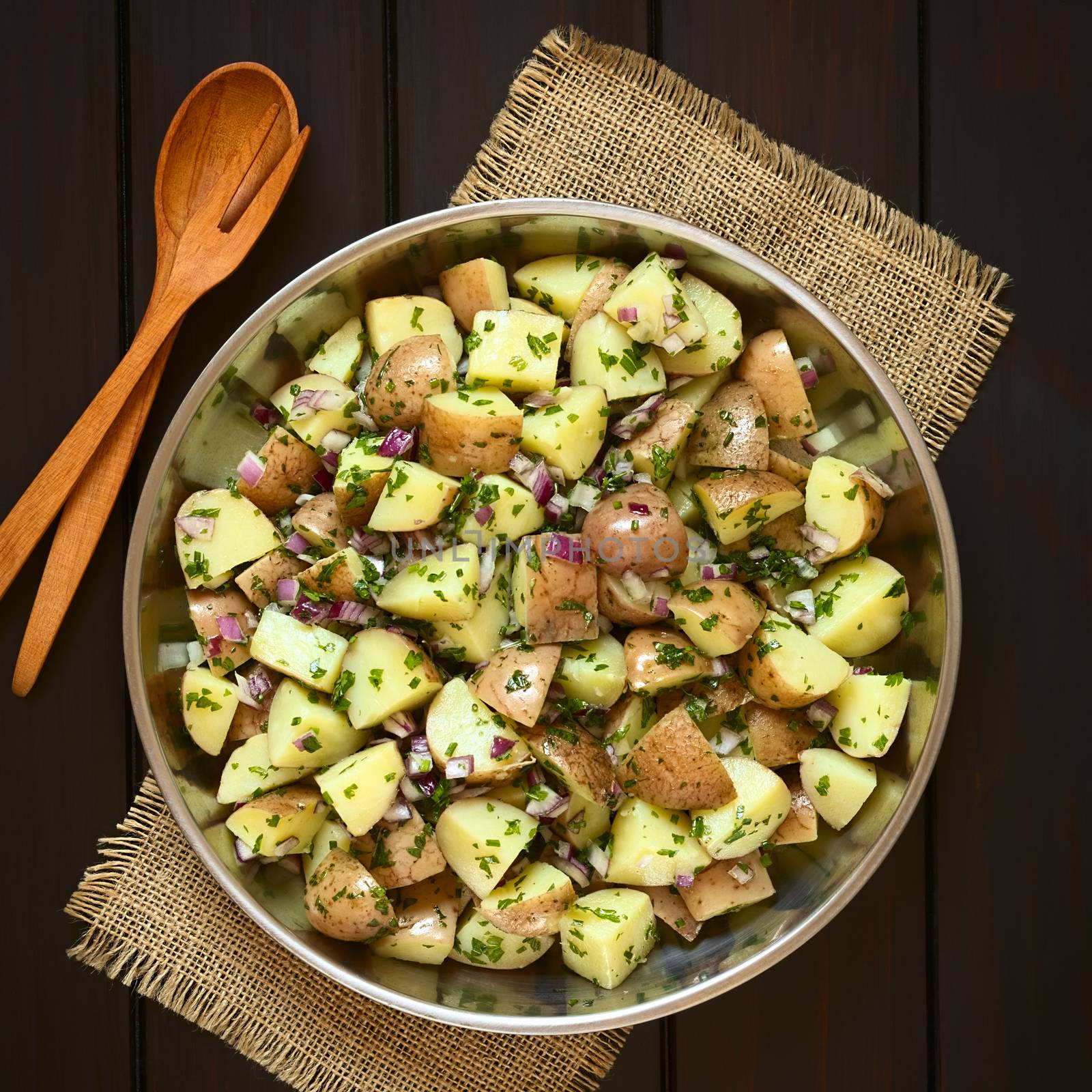 Salad of jacket potato, red onion and herbs in bowl with wooden spoon and fork, photographed overhead on dark wood with natural light