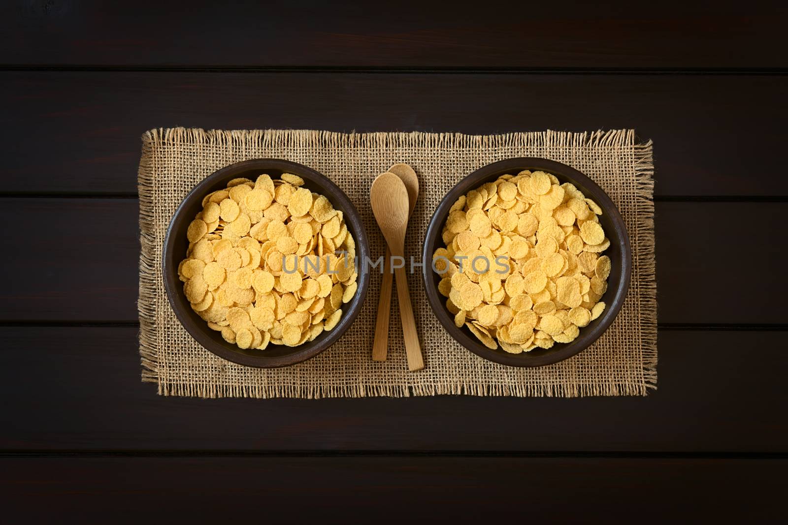 Crispy corn flakes breakfast cereal in rustic bowls with small wooden spoons, photographed overhead on dark wood with natural light