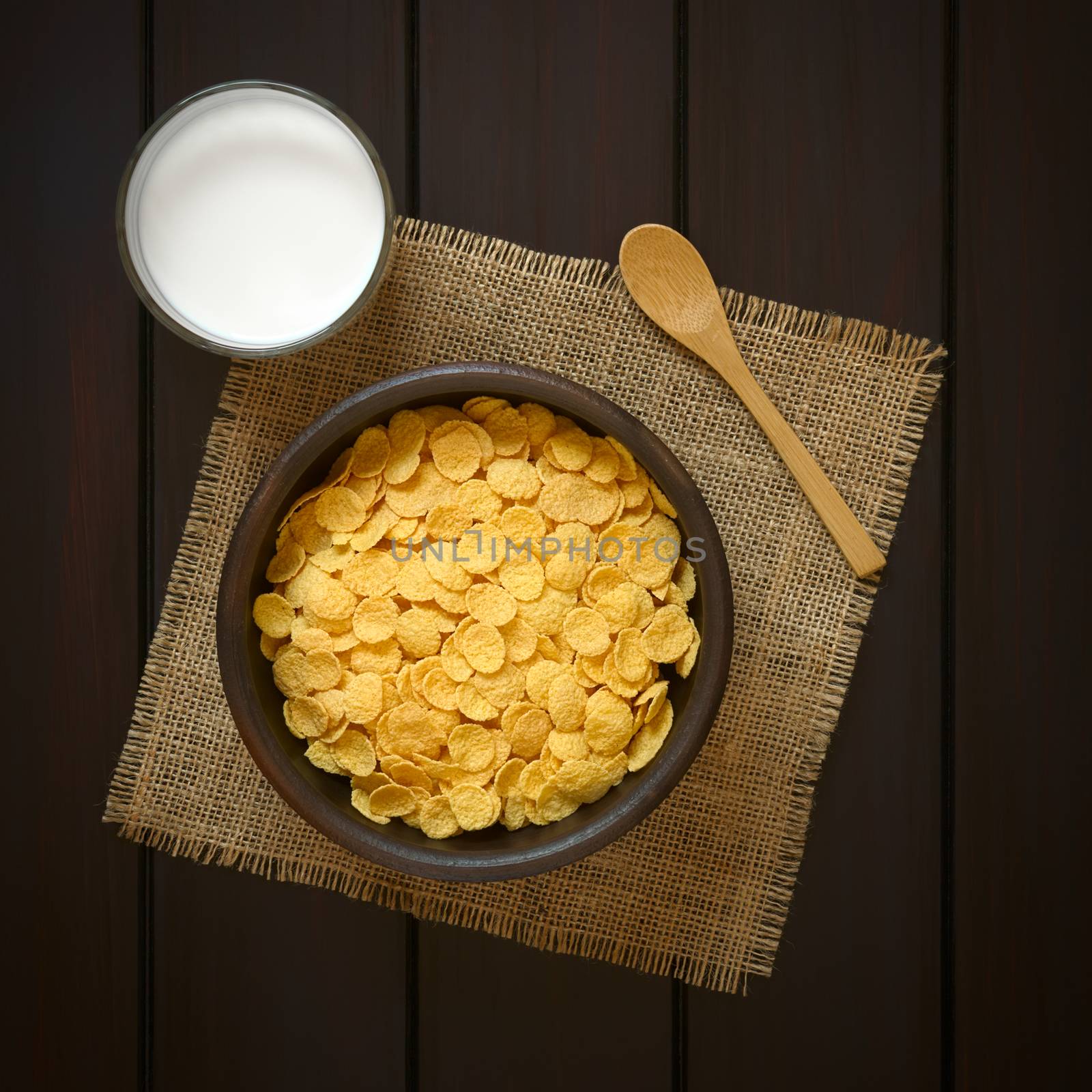 Crispy corn flakes breakfast cereal in rustic bowl with a glass of milk and a wooden spoon on the side, photographed overhead on dark wood with natural light