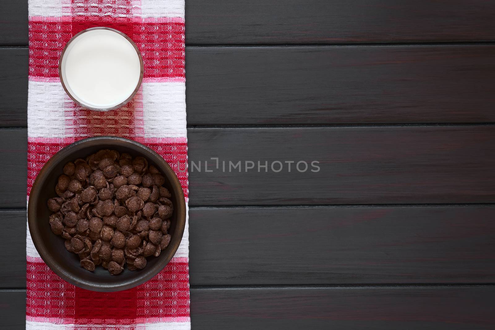 Crispy chocolate corn flakes breakfast cereal in rustic bowl with a glass of milk, photographed overhead on dark wood with natural light