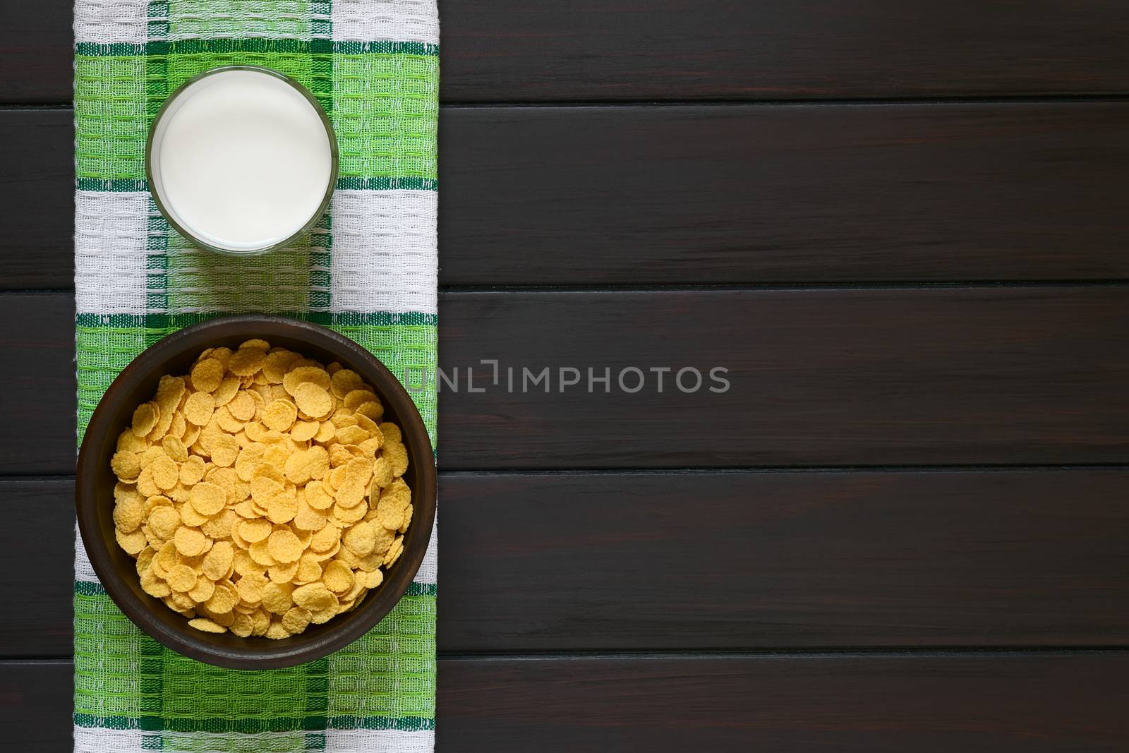 Crispy corn flakes breakfast cereal in rustic bowl with a glass of milk, photographed overhead on dark wood with natural light