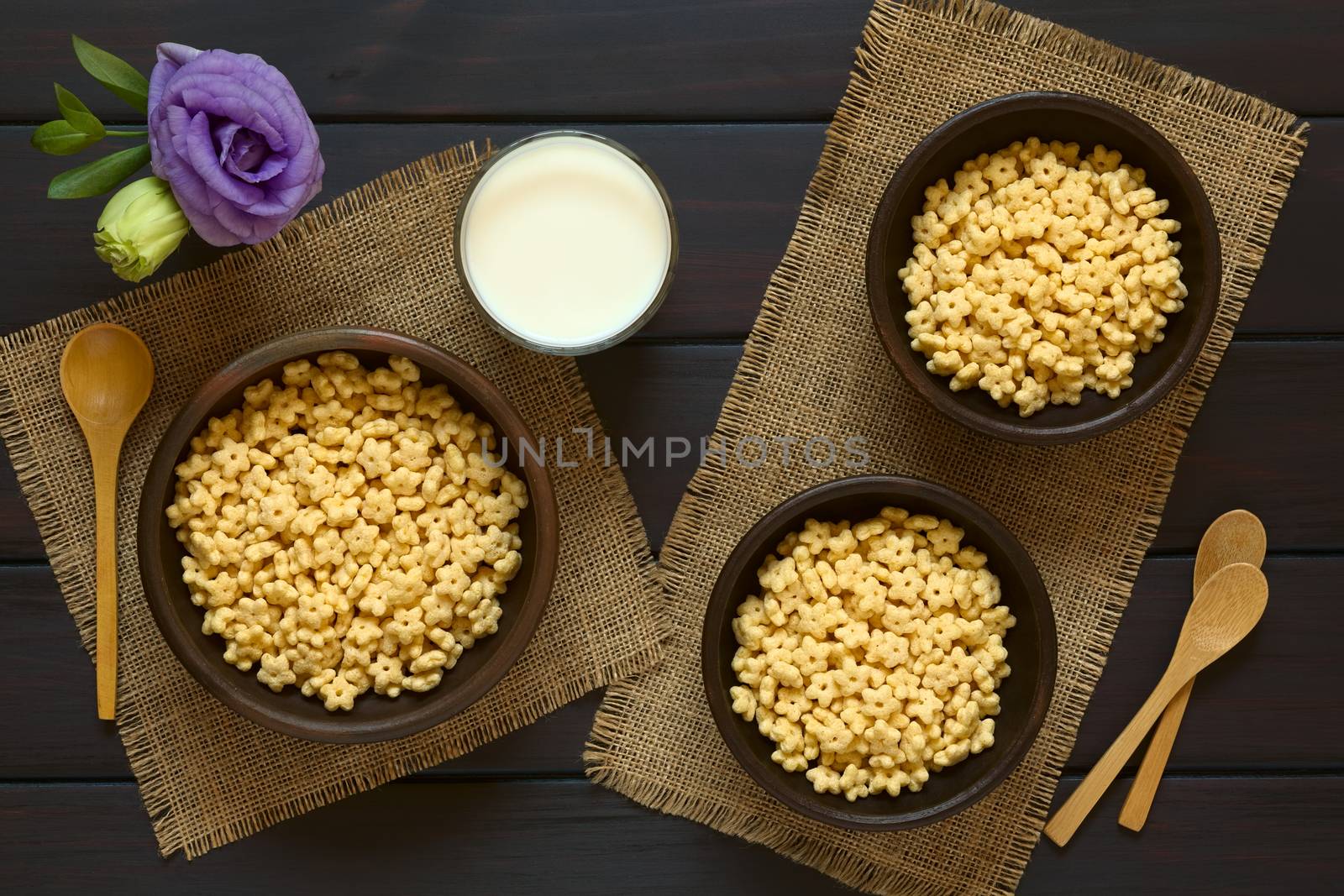 Honey flavored breakfast cereal in three rustic bowls with a glass of milk and wooden spoons on the side, photographed overhead on dark wood with natural light 
