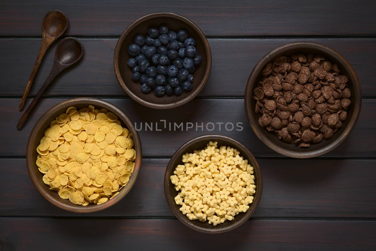 Crispy chocolate and simple corn flakes, and honey flavored breakfast cereal in rustic bowls with fresh blueberries and wooden spoons on the side, photographed overhead on dark wood with natural light