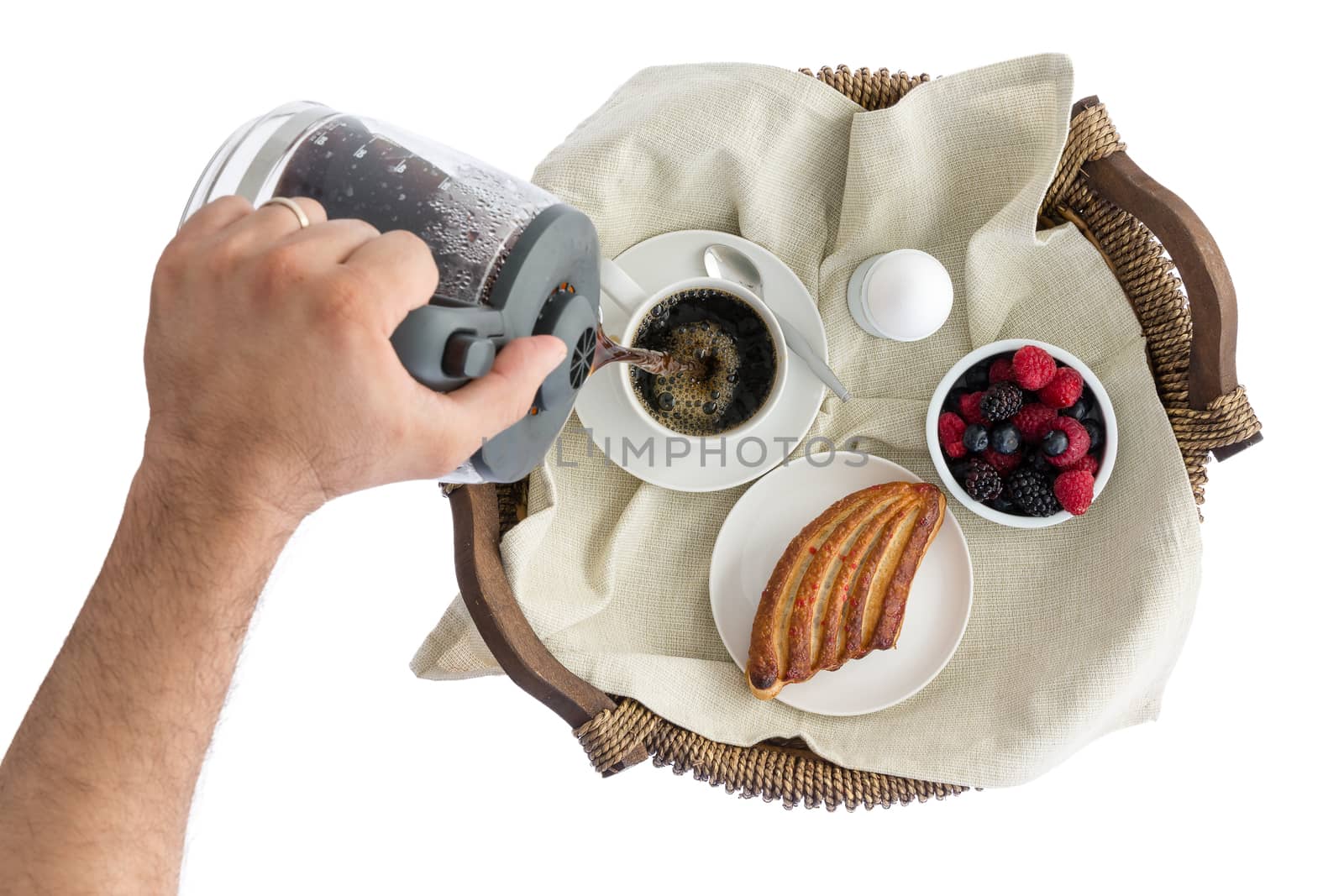 Overhead view of the hand of a man pouring himself fresh breakfast coffee in a cup on a breakfast tray with fresh berries, a pastry and boiled egg, isolated on white