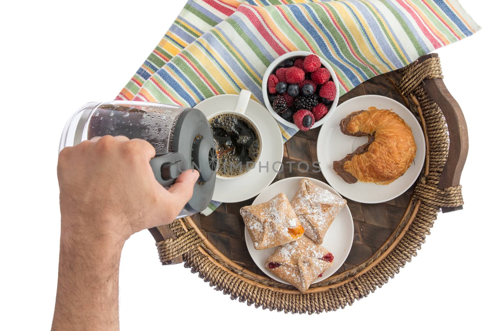 Man pouring fresh coffee for his breakfast from a glass jug into a cup on a wicker tray with assorted berries and pastries and a chocolate hazelnut croissant, overhead on white