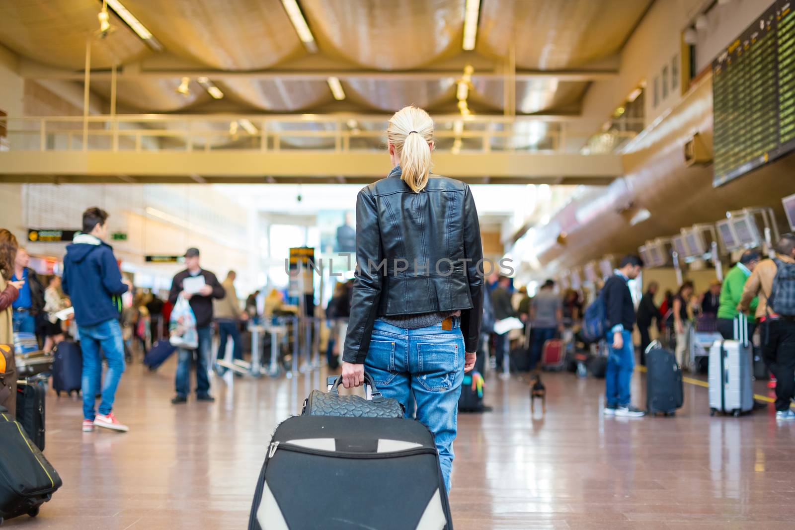 Casually dressed young stylish female traveller walking the airport terminal hall   draging suitcase and a handbag behind her. Blured background. Can also be used as railway, metro, bus station.