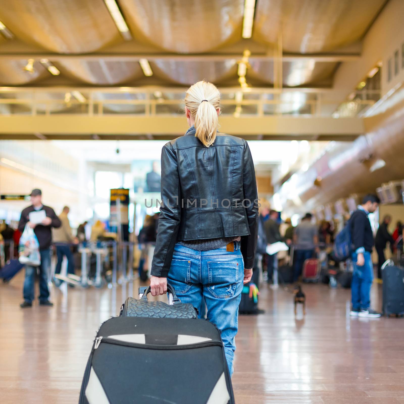 Casually dressed young stylish female traveller walking the airport terminal hall   draging suitcase and a handbag behind her. Blured background. Can also be used as railway, metro, bus station.