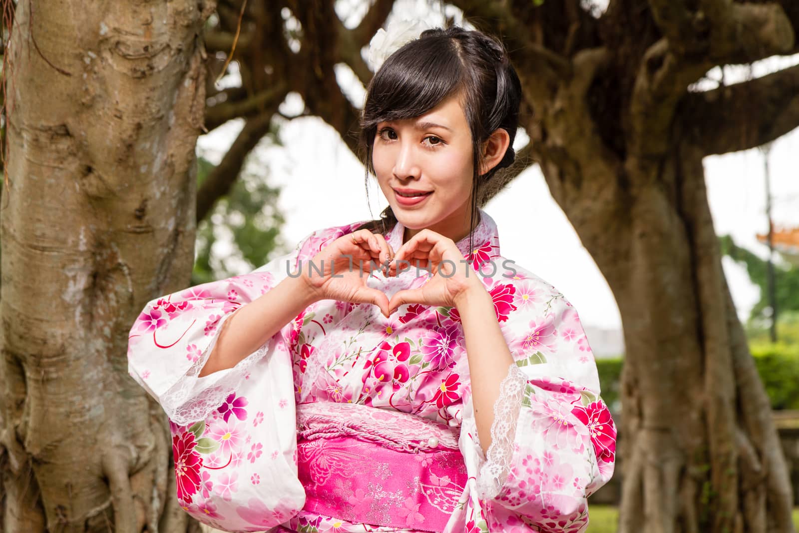 Asian woman in kimono by trees in garden, making a heart with her hands