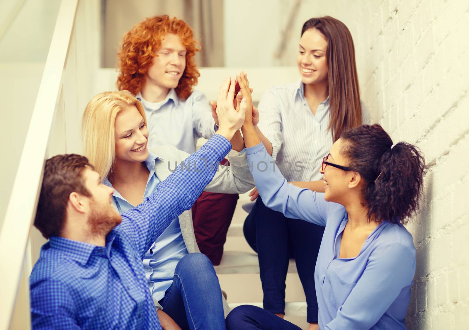 business, office, gesture and startup concept - smiling creative team doing high five gesture sitting on staircase