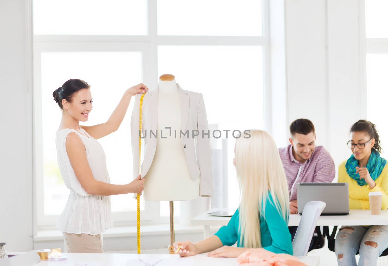 startup, education, fashion and office concept - smiling designers drawing sketches and measuring jacket on mannequin in office