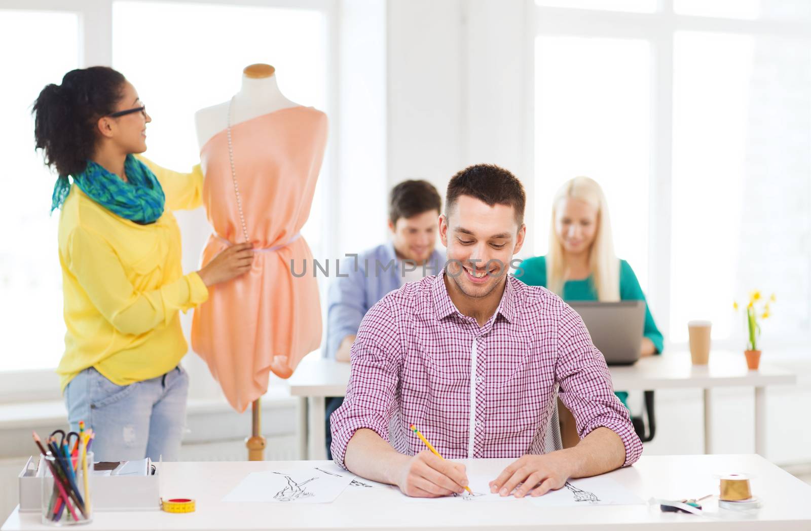 startup, education, fashion and office concept - smiling male drawing sketches and female adjusting dress on mannequin in office