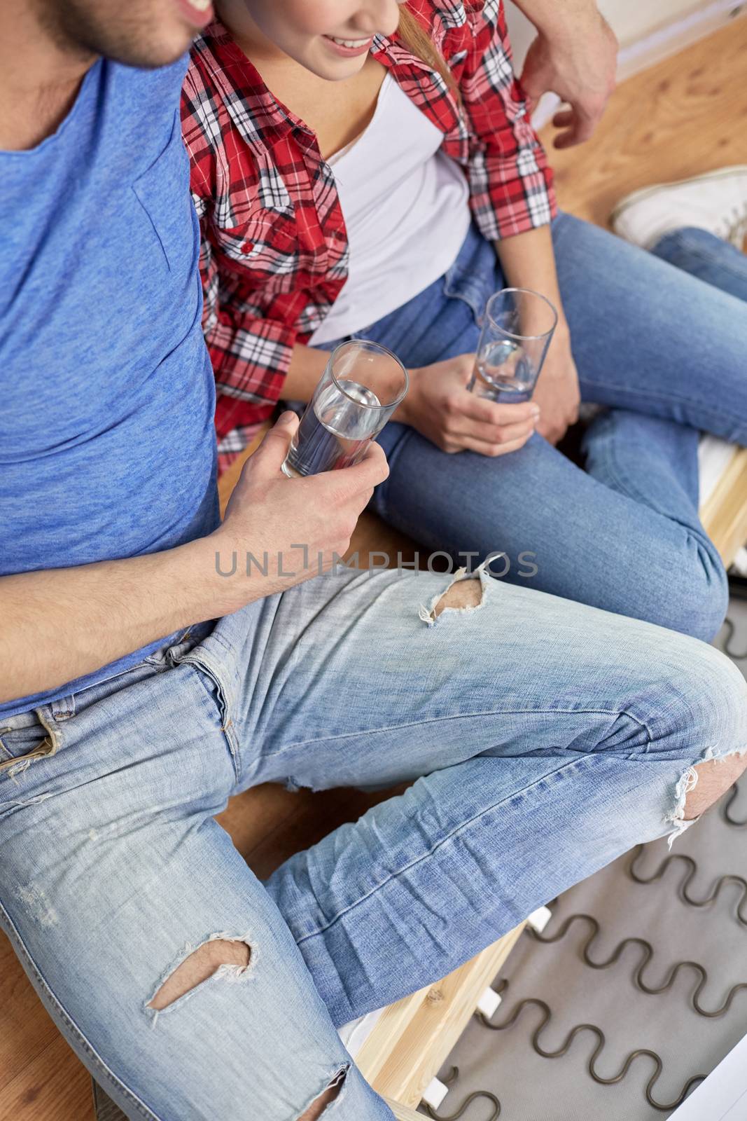 moving, home, repair and people concept - close up of couple legs relaxing and and drinking water from glasses in new apartment