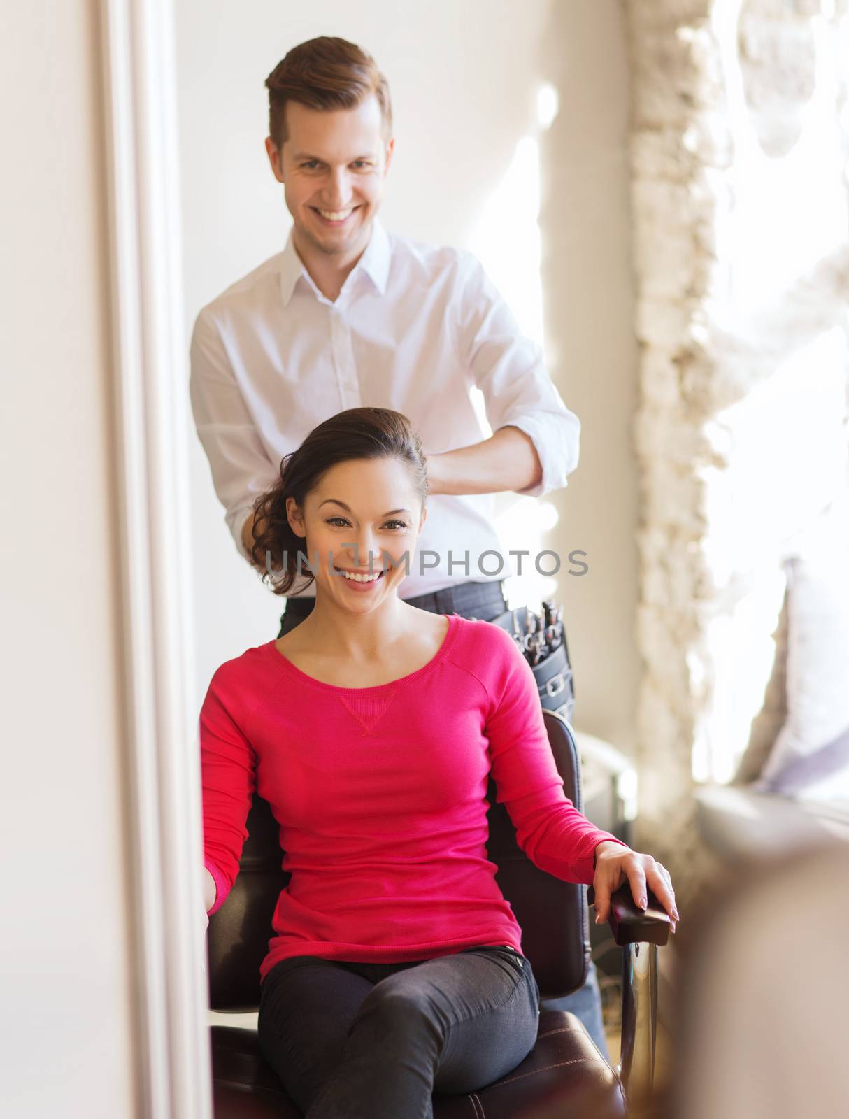 beauty, hairstyle and people concept - happy young woman with hairdresser finishing hairdo at salon