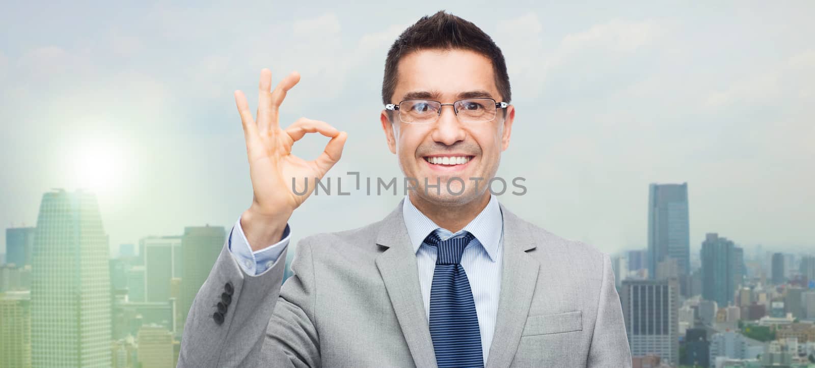 business, people, vision and success concept - happy smiling businessman in eyeglasses and suit showing ok sign over city background
