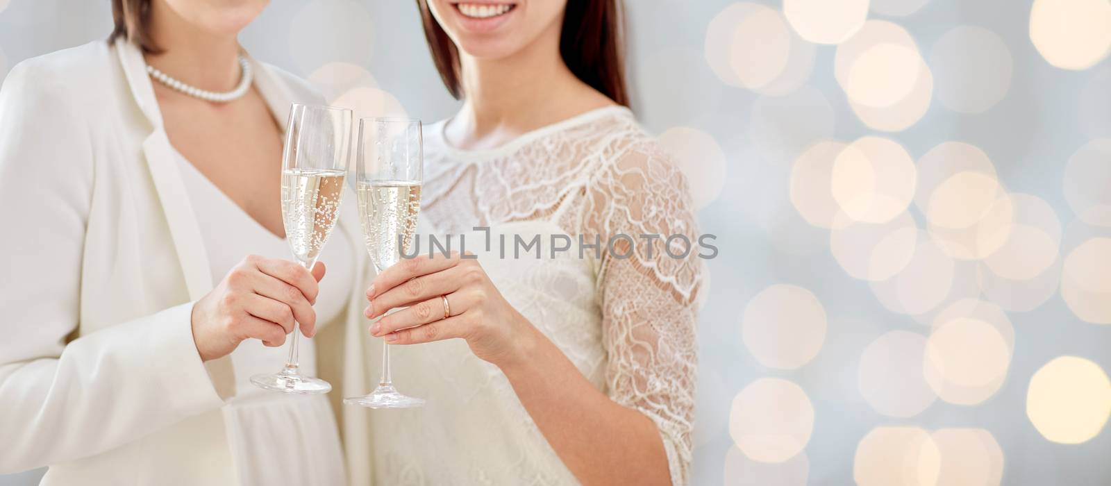 people, homosexuality, same-sex marriage, celebration and love concept - close up of happy married lesbian couple holding and clinking champagne glasses over holidays lights background